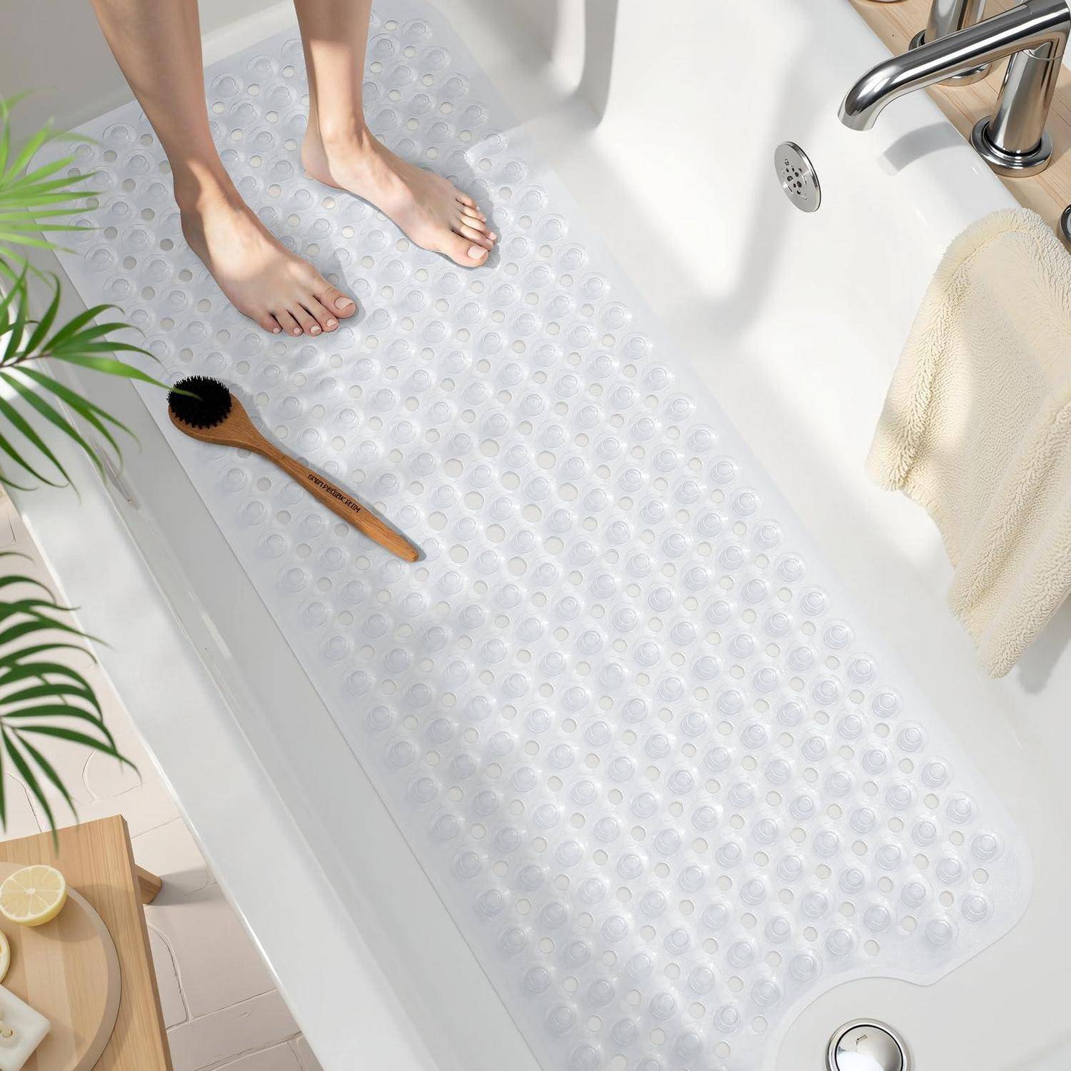 Füße, die auf einer durchsichtigen Badematte mit Saugnäpfen in einer weißen Badewanne stehen, mit einer Holzbürste, einem Handtuch und einem Wasserhahn in der Nähe.