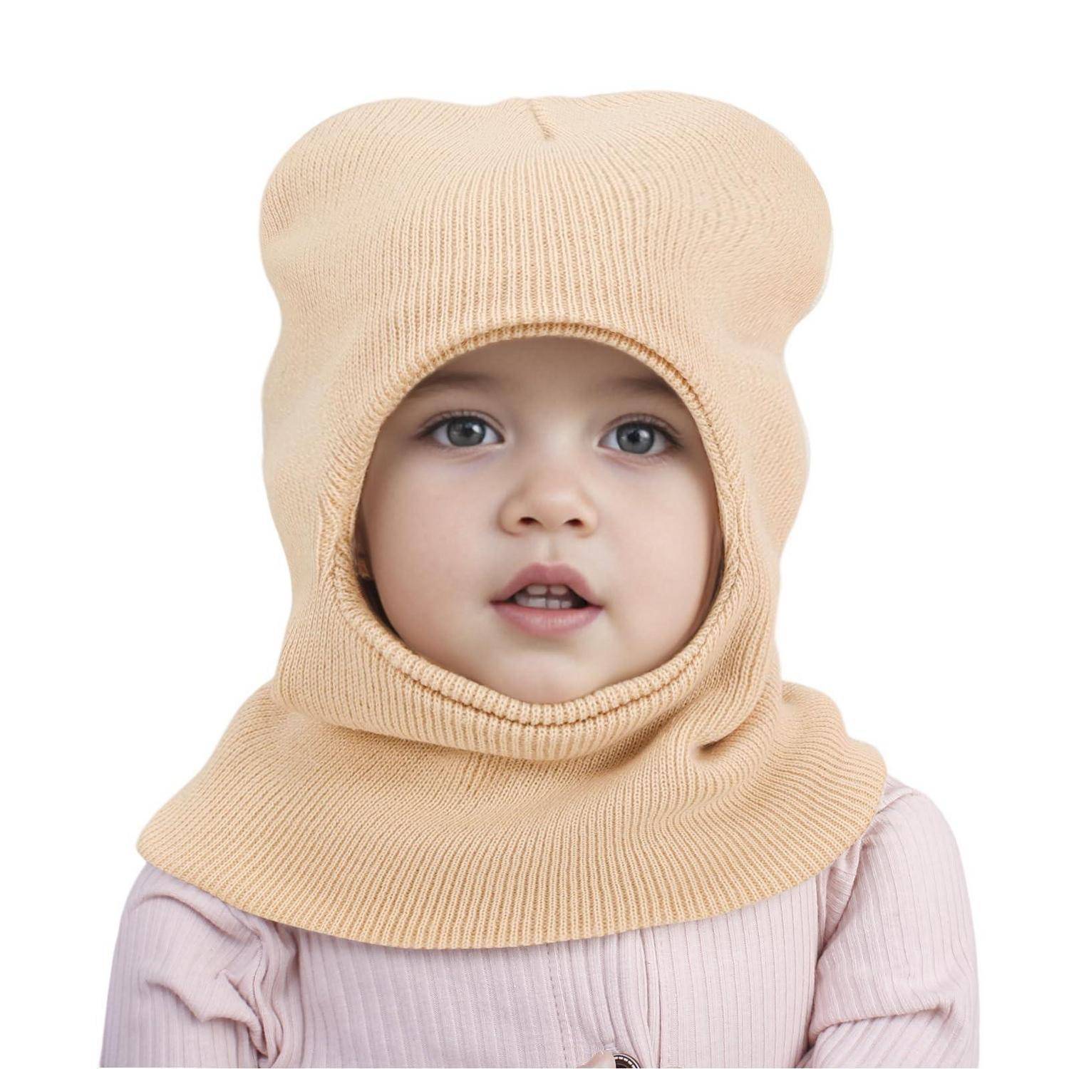 Porträt eines Babys mit einer beigefarbenen gestrickten Sturmhaube mit Ohrenschutzen vor einem einfachen weißen Hintergrund.