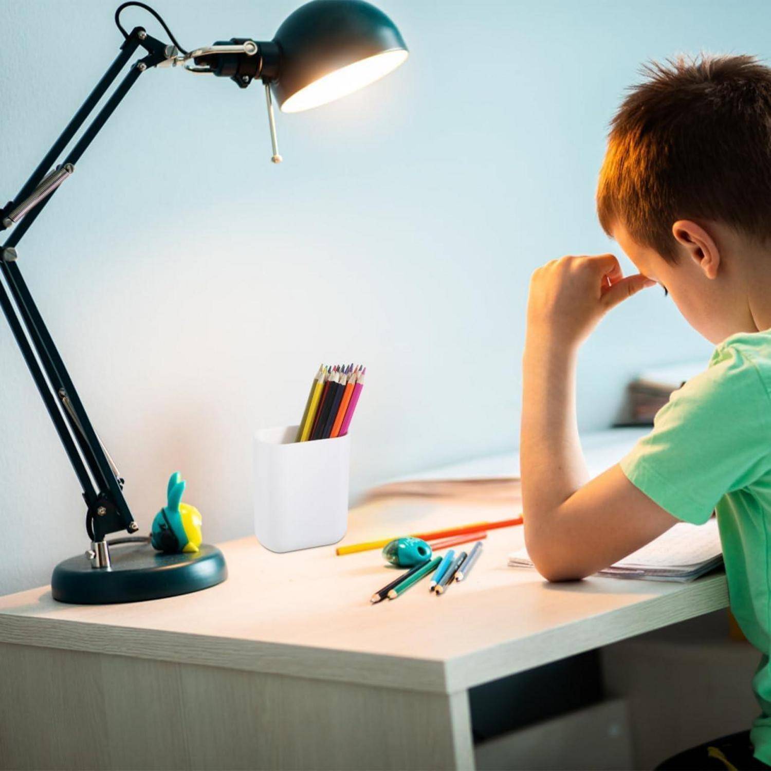 Ein Junge sitzt an einem Schreibtisch unter einer brennenden Lampe und lernt, umgeben von Stiften und Notizbüchern. Die Szene wird von hinten fotografiert in einem ruhigen, häuslichen Arbeitsbereich.