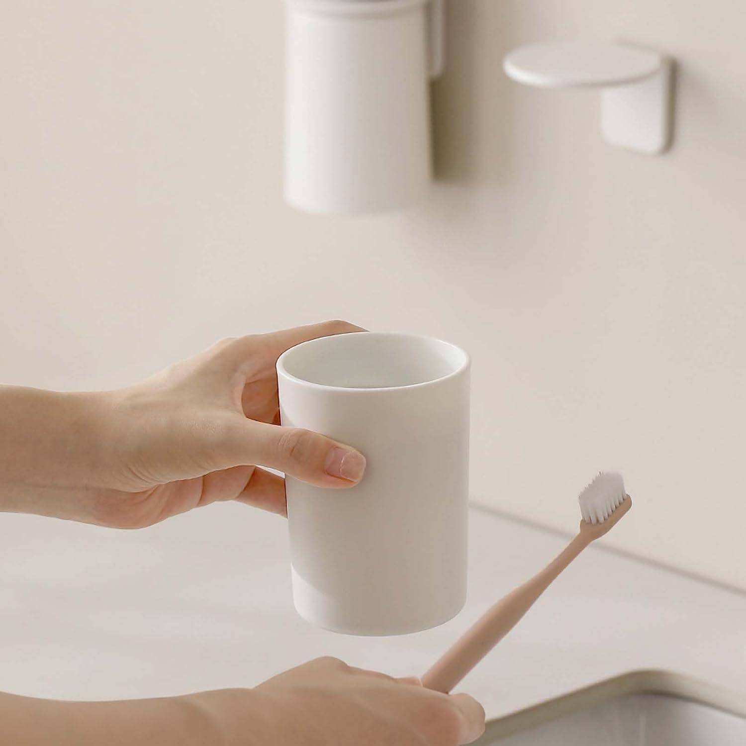Hand über einem weißen Becher über einem Badezimmerspülbecken, mit einer Zahnbürste in der anderen Hand und einem wandmontierten Becherhalter oben.