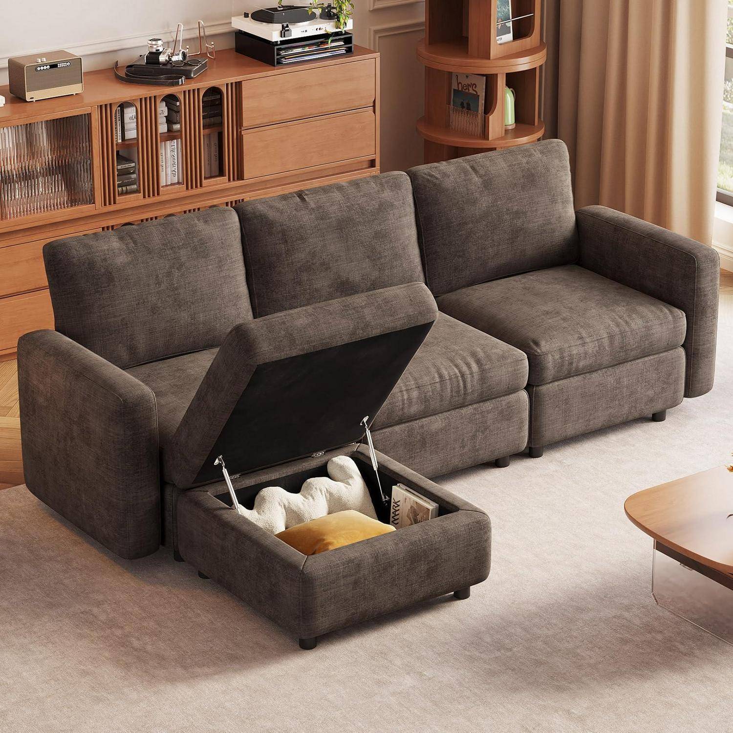 Grau gepolstertes Modulsofa mit einer aufklappbaren Stauraum-Ottomane, die Decken und Bücher enthält, in einem modernen Wohnzimmer.