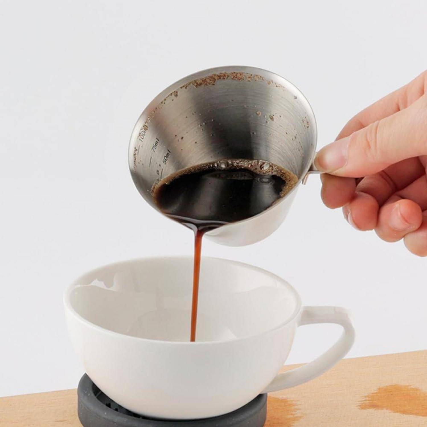 Eine Hand gießt Kaffee durch einen Metallfilter in eine weiße Tasse auf einer Untertasse, wobei etwas Kaffee auf dem Tisch verschüttet wird.