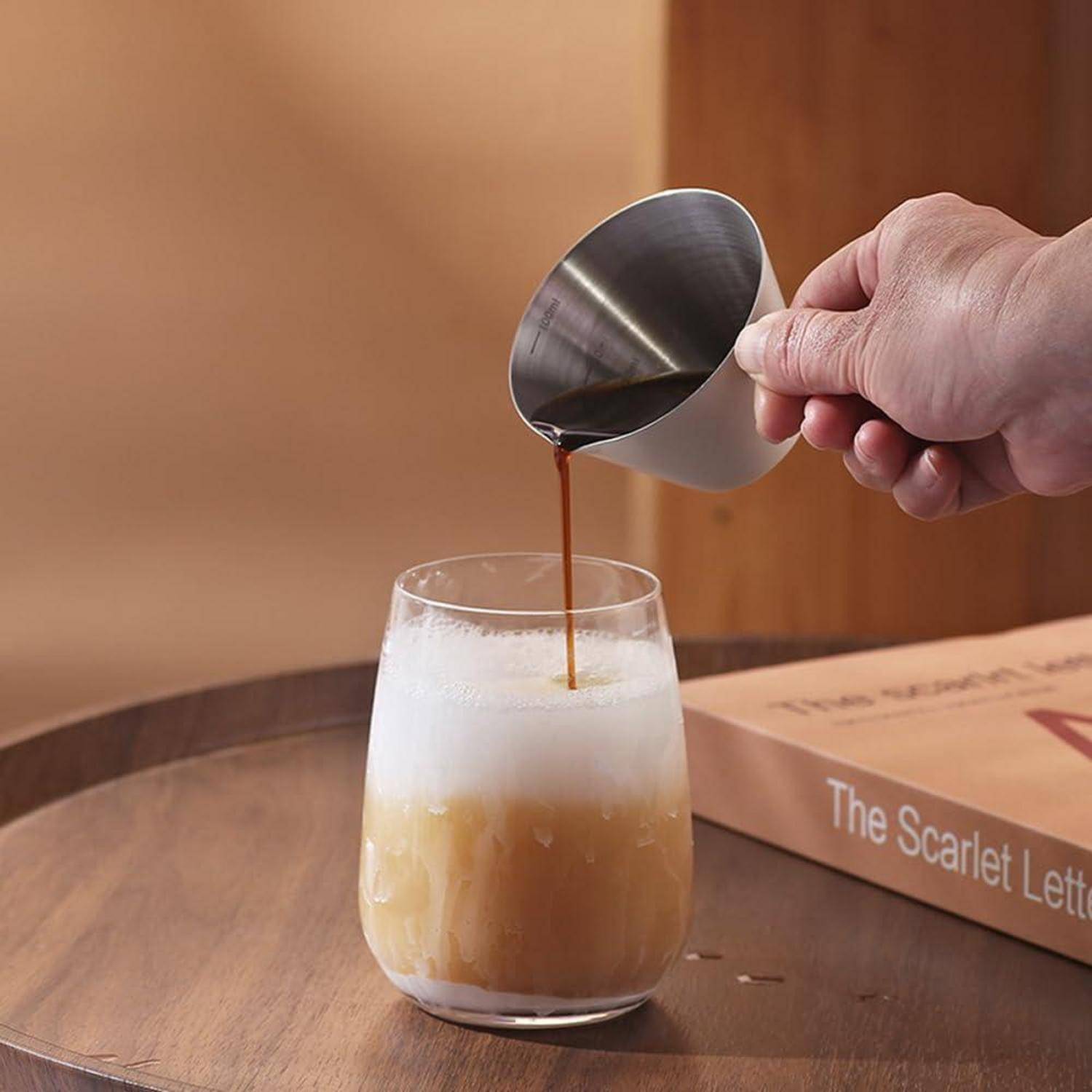 Eine Hand gießt Kaffee in ein schäumiges Eiskaltgetränk in ein Glas auf einem Holztisch neben einem Buch mit dem Titel „The Scarlet Letter