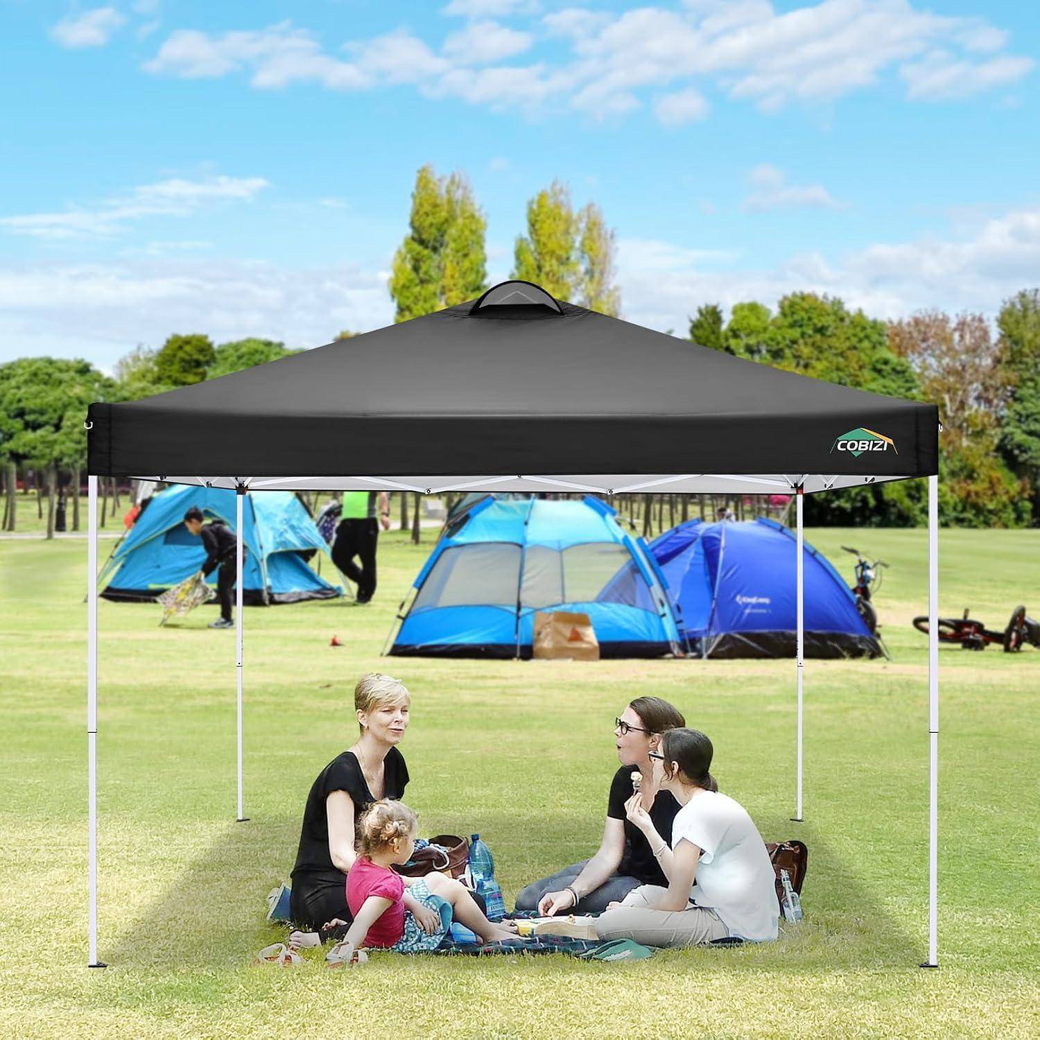 Familie, die auf einer Picknickdecke unter einem schwarzen Pavillon im Park sitzt, mit Campingzelten im Hintergrund.
