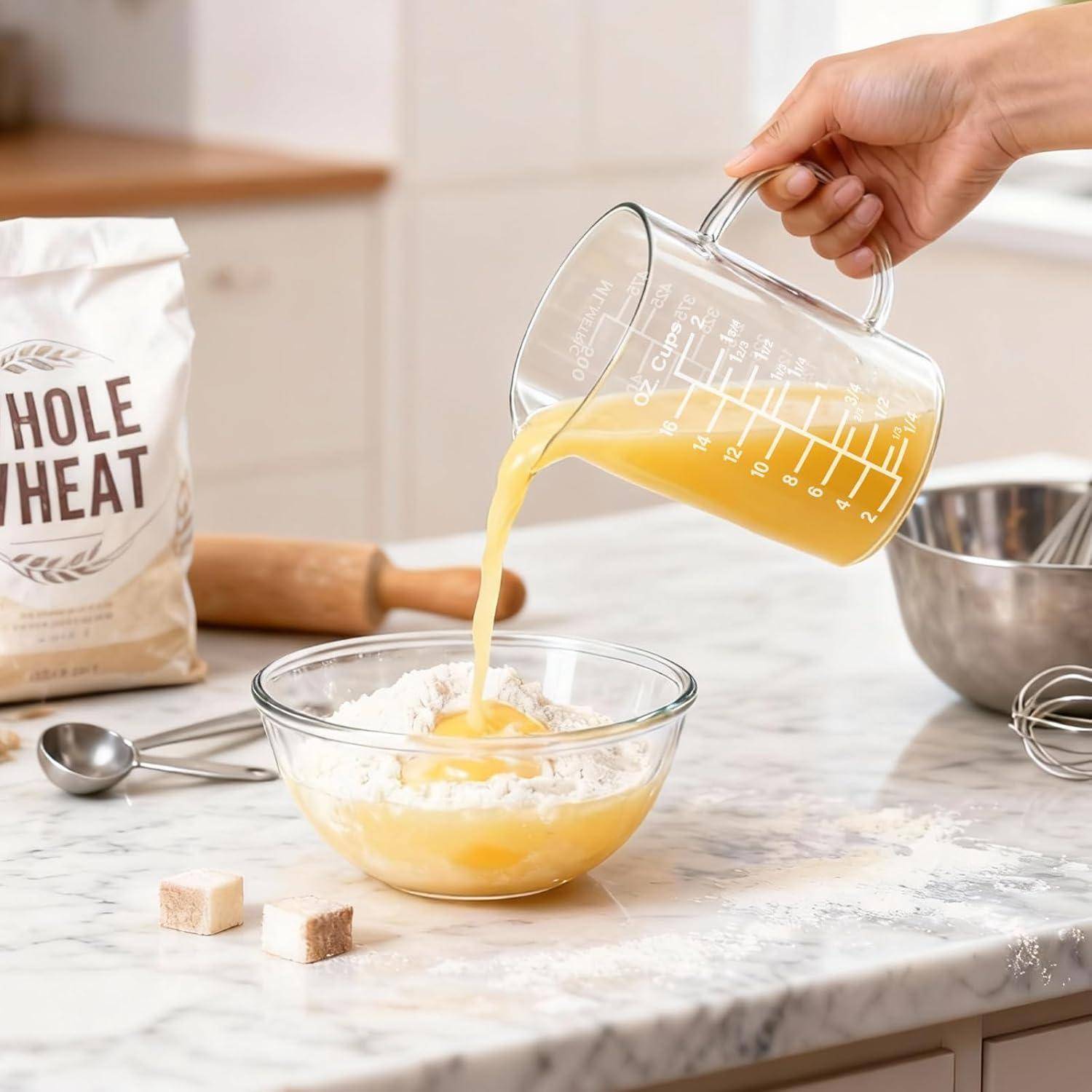 Eine Hand gießt geschlagene Eier aus einem Glaskmeßbecher in eine Schüssel mit Mehl auf einer Küchenarbeitsplatte, mit Vollkornmehl und Backwerkzeugen in der Nähe.