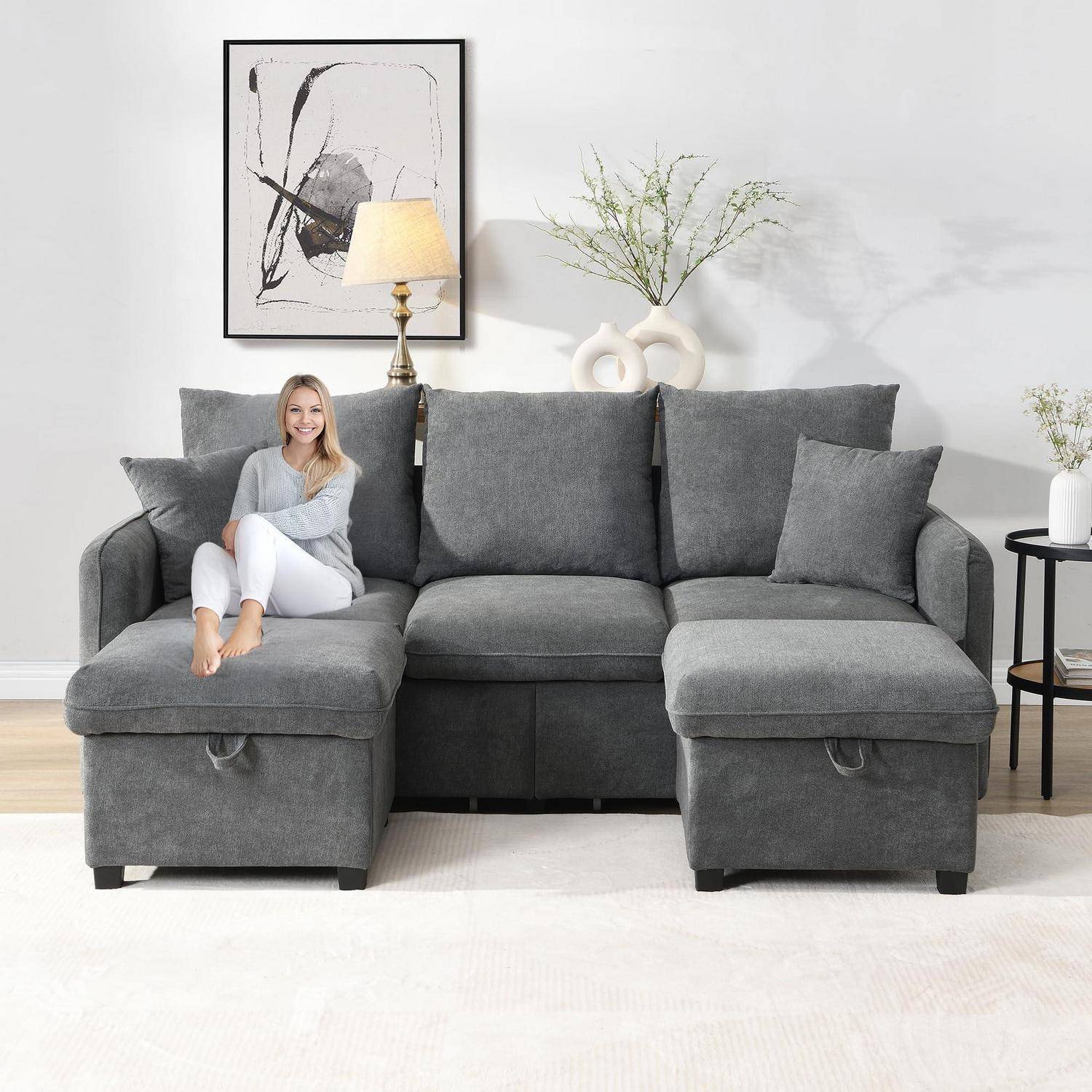 Graue U-förmige Ecksofa mit Chaiselongue; eine Frau sitzt auf der linken Chaiselongue in einem hellen Wohnzimmer mit Lampe, Wandkunst und Dekoration.