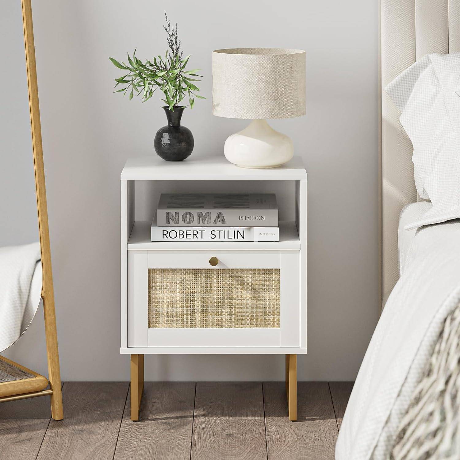 Weißer Nachttisch mit Lampe, Vase mit Grünpflanzen und Büchern neben einem Bett in einem hellen Schlafzimmer.