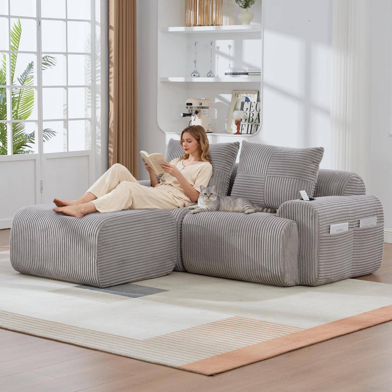 Frau, die auf einem grau gestreiften Ecksofa mit einer Katze liest, in einem hellen Wohnzimmer mit Regalen und einem großen Teppich.