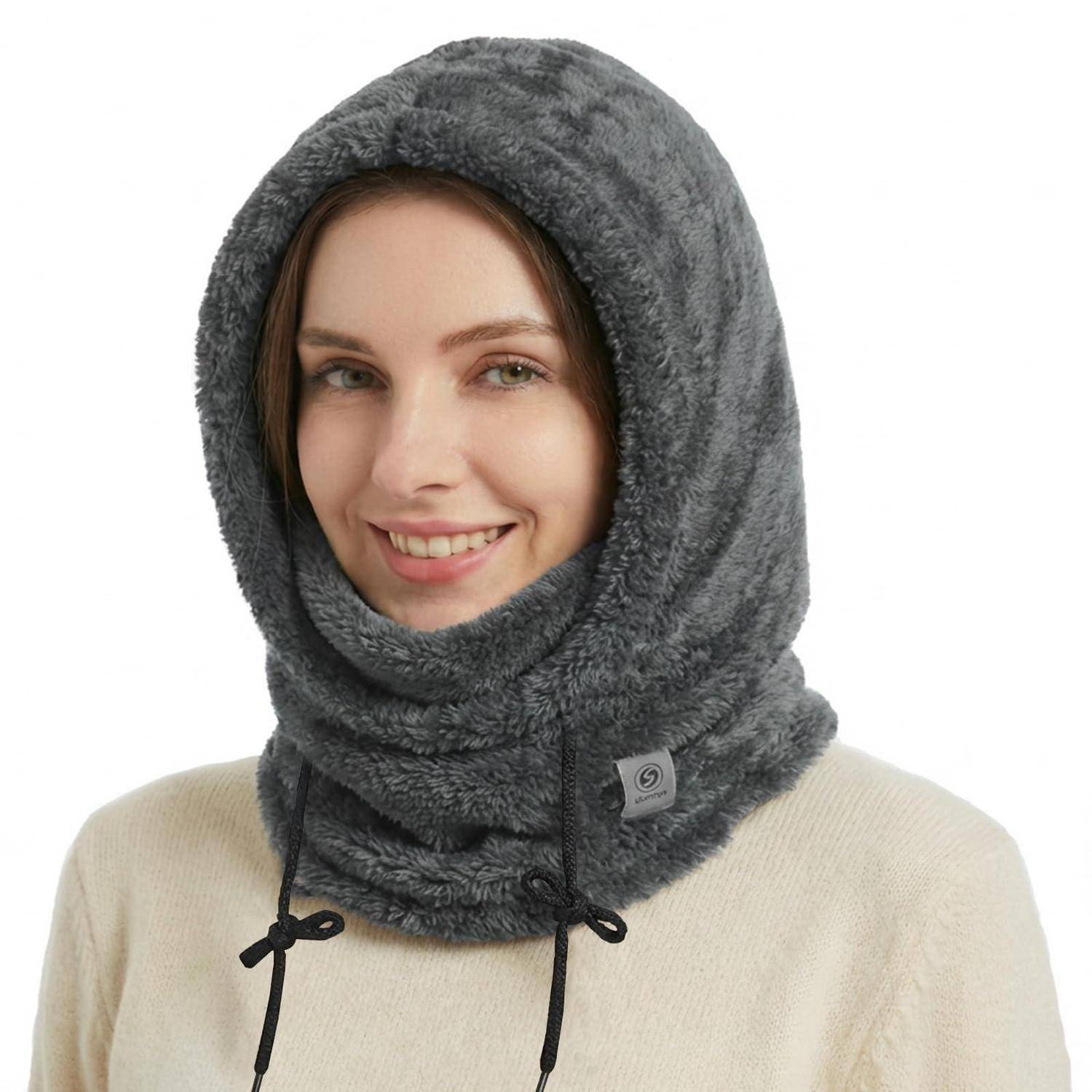 Lächelnde Frau trägt eine graue Fleece-Sturmhaube mit Kapuze und Zugschnüren über einem beigen Pullover vor weißem Hintergrund.