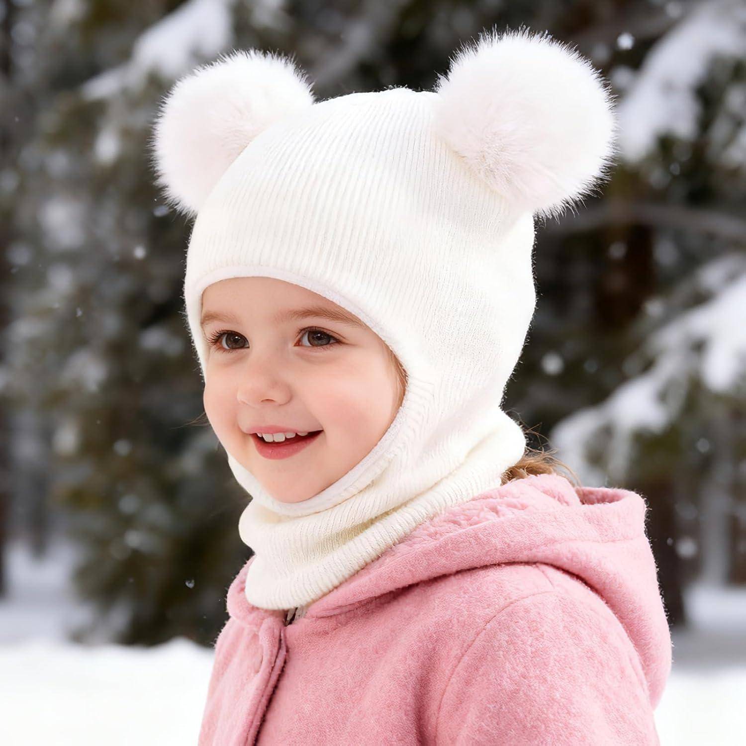 Lachendes kleines Kind im Freien im Schnee, das eine weiße Strickmütze mit Pompon-Ohren und einen rosa Mantel trägt.