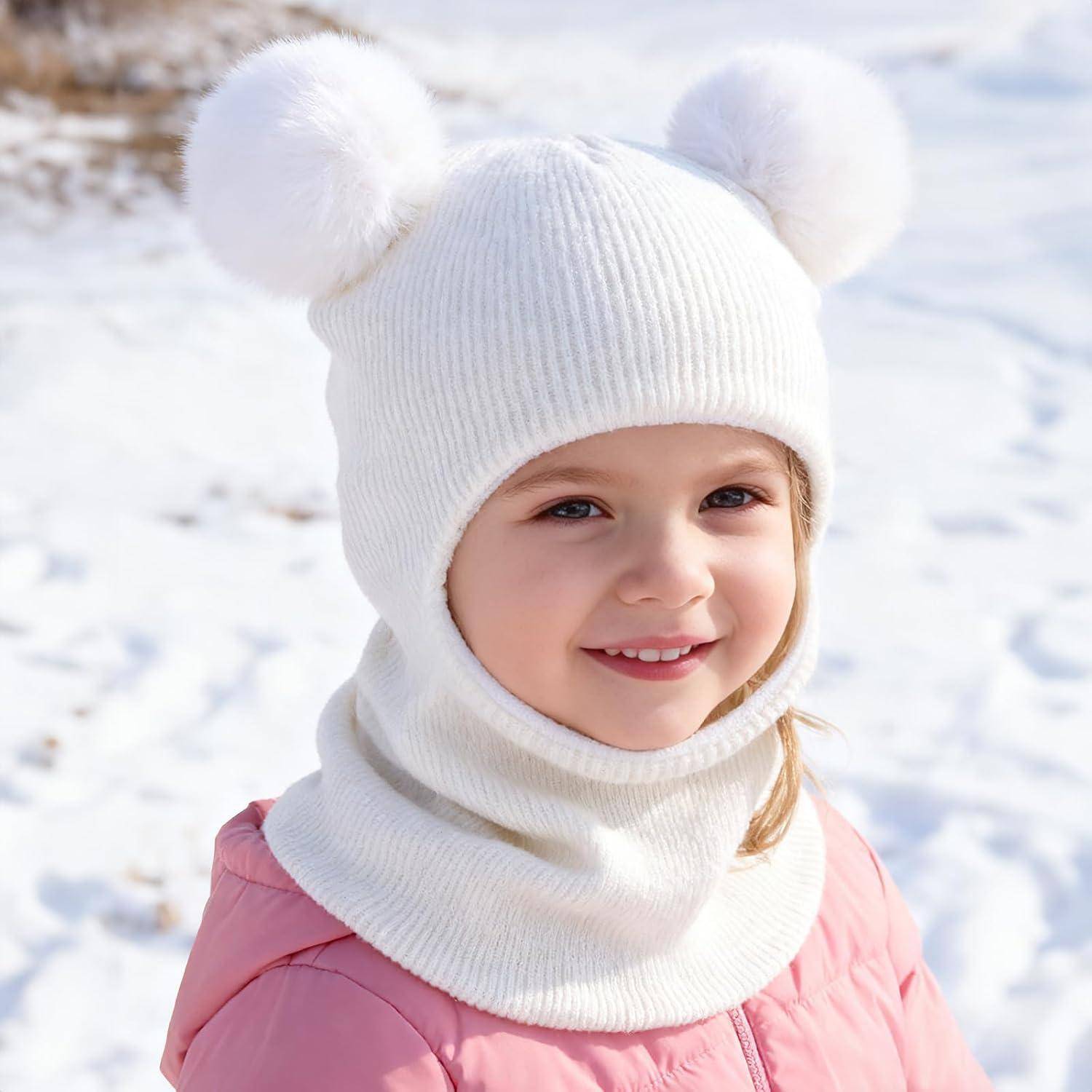 Lächelndes Kind in einer weißen Wintermütze mit Bommelchen und passendem Schal, das einen rosa Mantel trägt und draußen im Schnee steht.