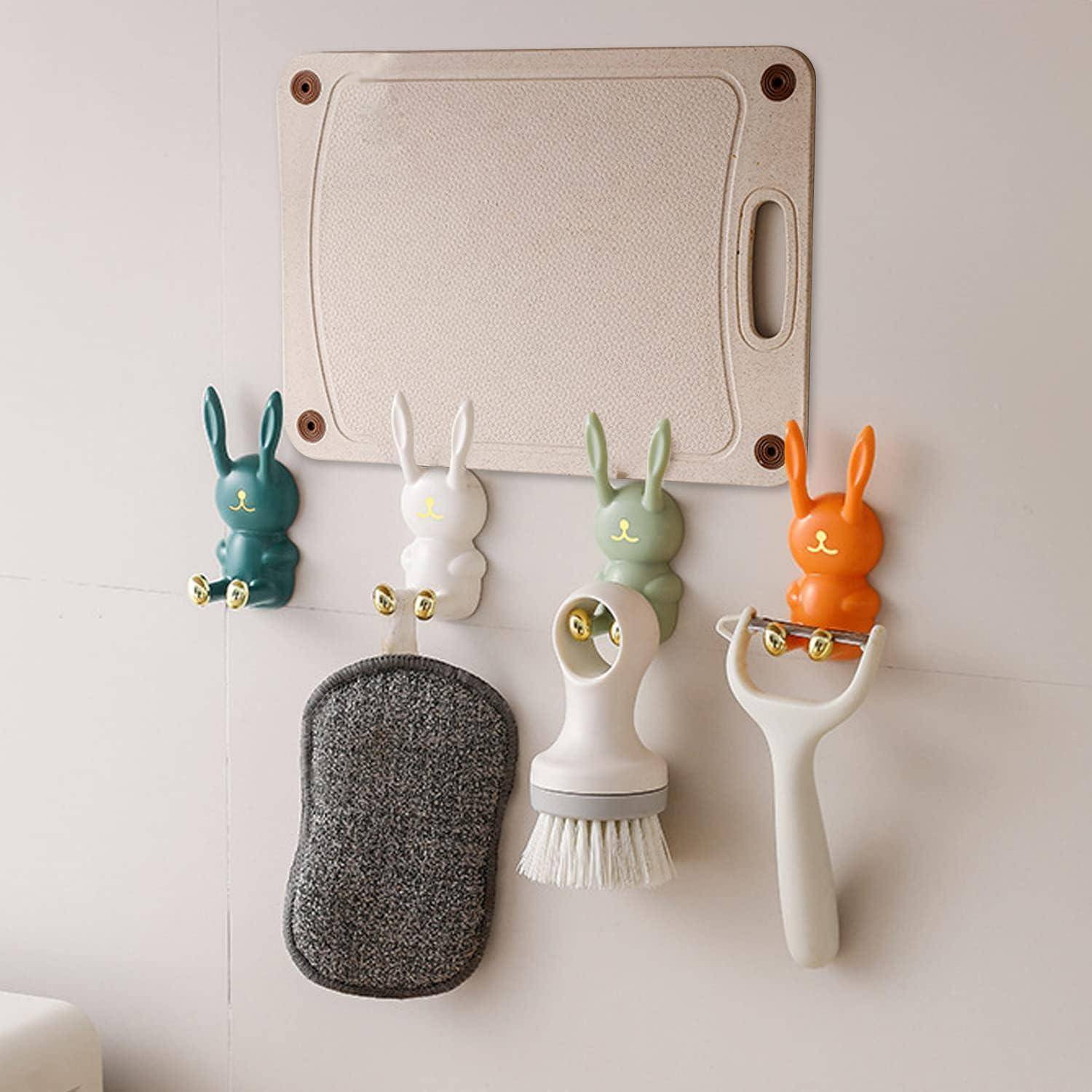 Wandmontierter Küchenorganizer mit hasenförmigen Haken, an denen ein Schwamm, eine Schrubbürste und ein Sparschäler hängen, unter einem kleinen Schneidebrett.