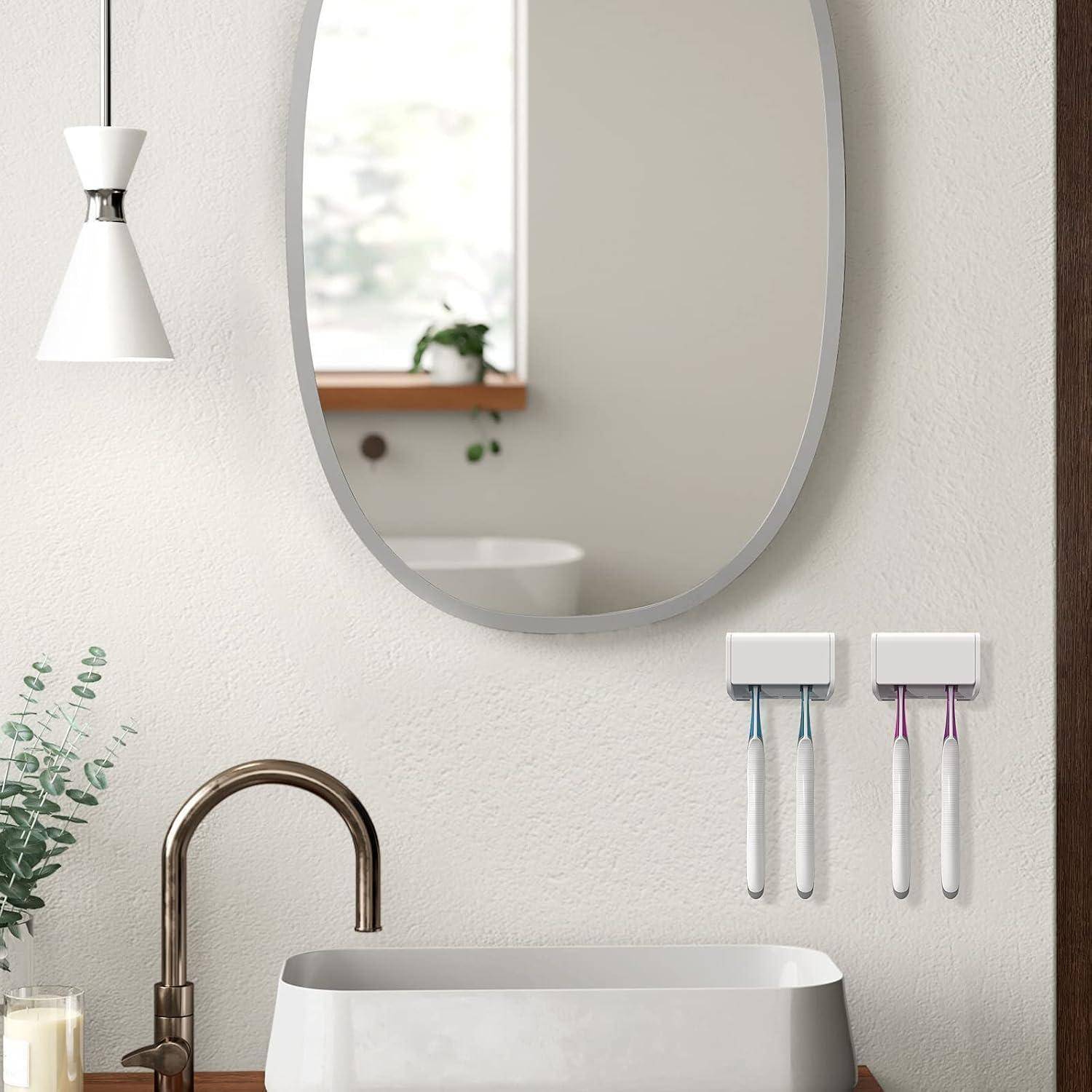 Modernes Badezimmer-Waschtisch mit ovalem Spiegel, Wandleuchte, Waschbecken-Armatur und montierte Zahnbürsterhalter in minimalistischem weißem Interieur.