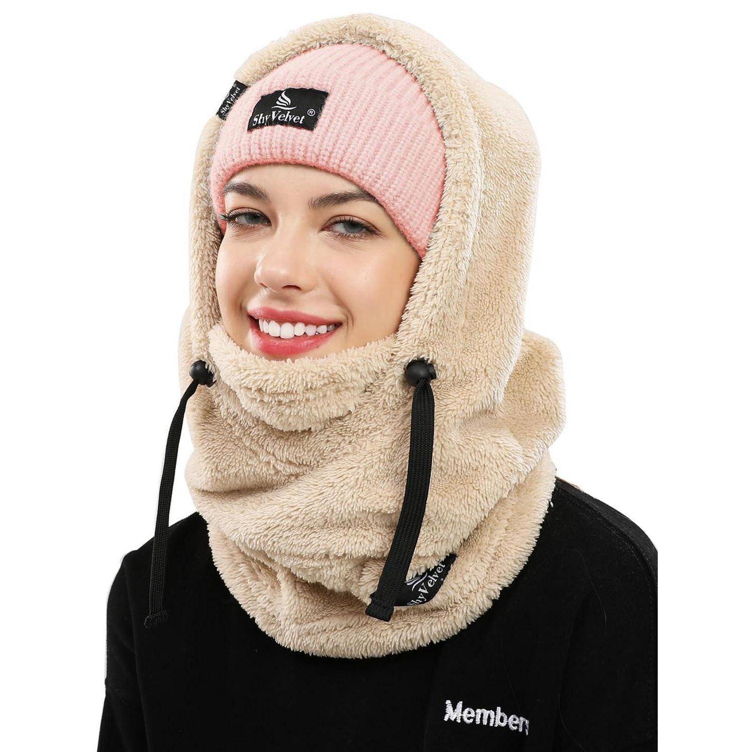 Lächelnde Frau, die eine beige Fleece-Sturmhaube und eine rosa Strickmütze mit schwarzen Zugbändern und einer schwarzen Jacke trägt, auf weißem Hintergrund.