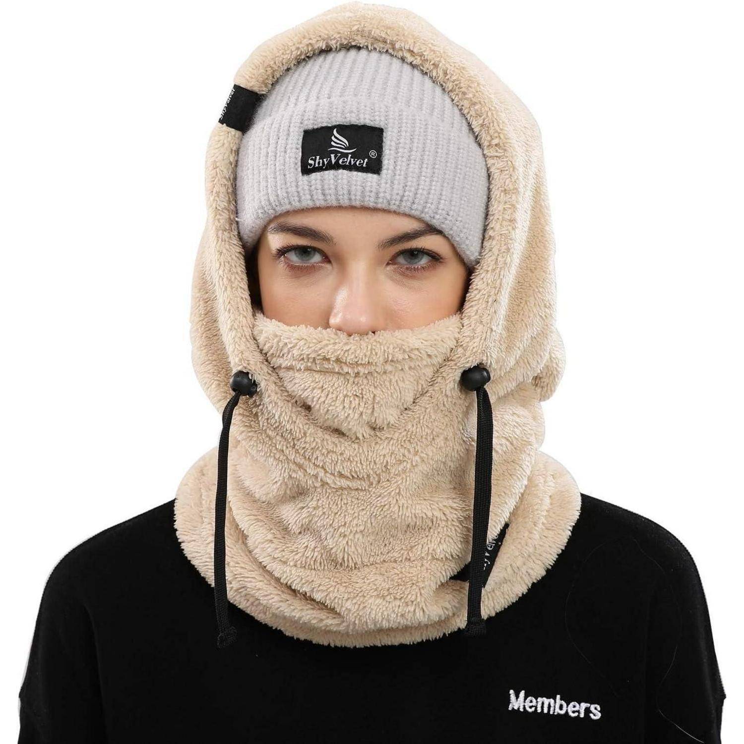 Person trägt eine beige Fleece-Sturmhaube und eine graue Mütze, die das Gesicht verdecken, wobei nur die Augen sichtbar sind, über einem schwarzen „Members"-Shirt auf weißem Hintergrund.