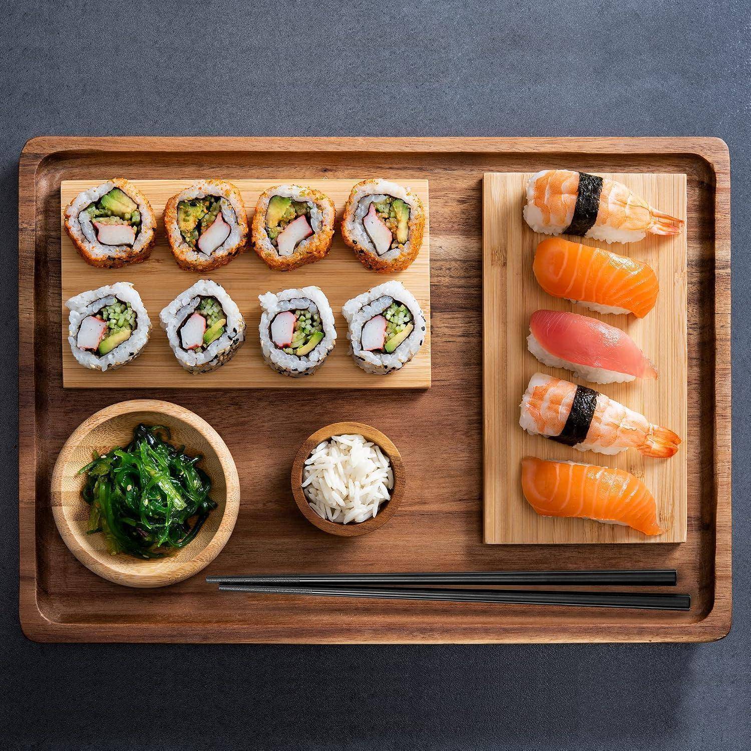 Holzplatte mit assortiertem Sushi: Maki-Rollen, Nigiri, Algensalat, eingelegter Ingwer und Stäbchen auf einem dunklen Tisch.
