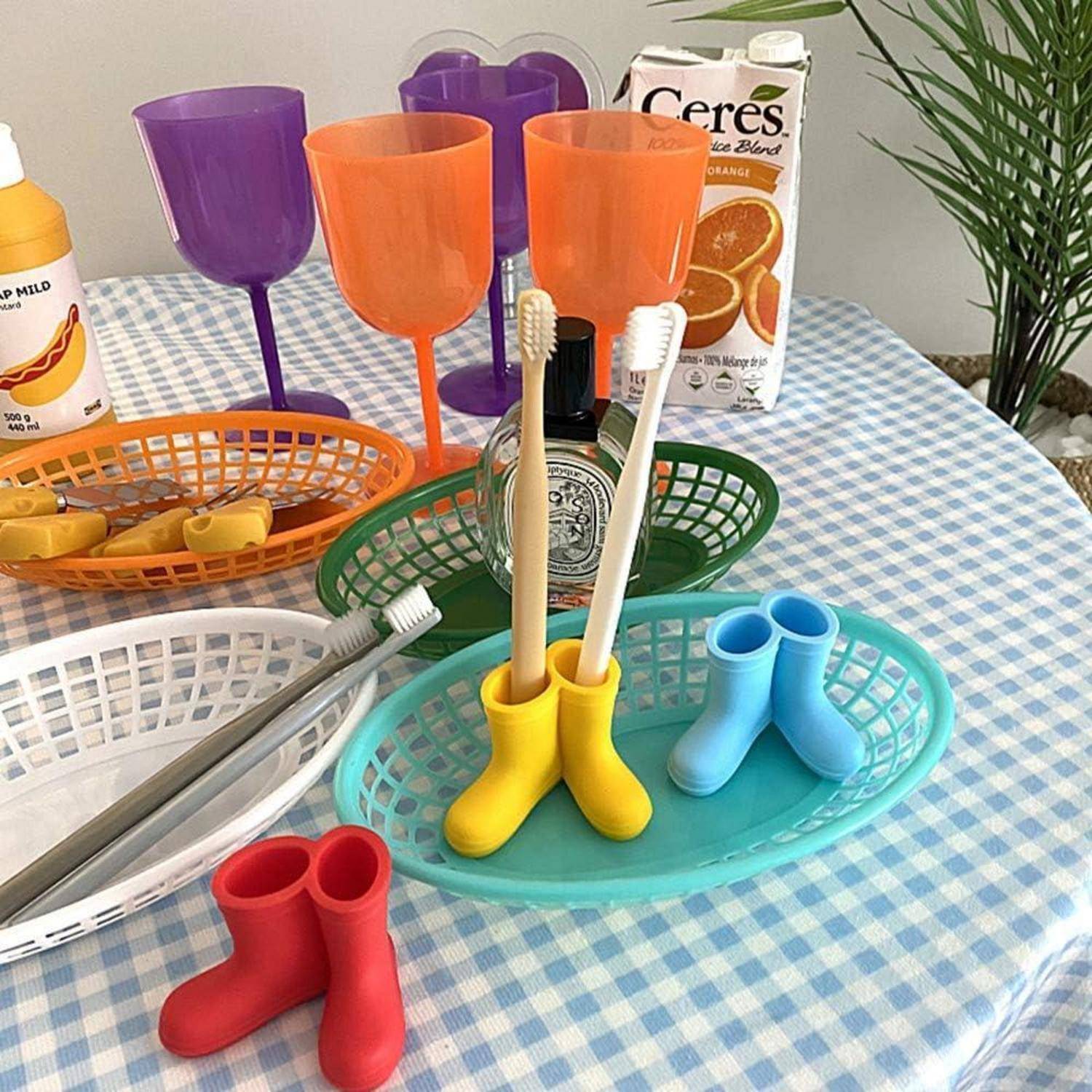 Bunte Kunststoffbecher, Körbe und stiefelförmige Zahnbürstenhalter auf einem karierten Tisch angeordnet, mit Seife und einem Orangensaftkarton im Hintergrund.