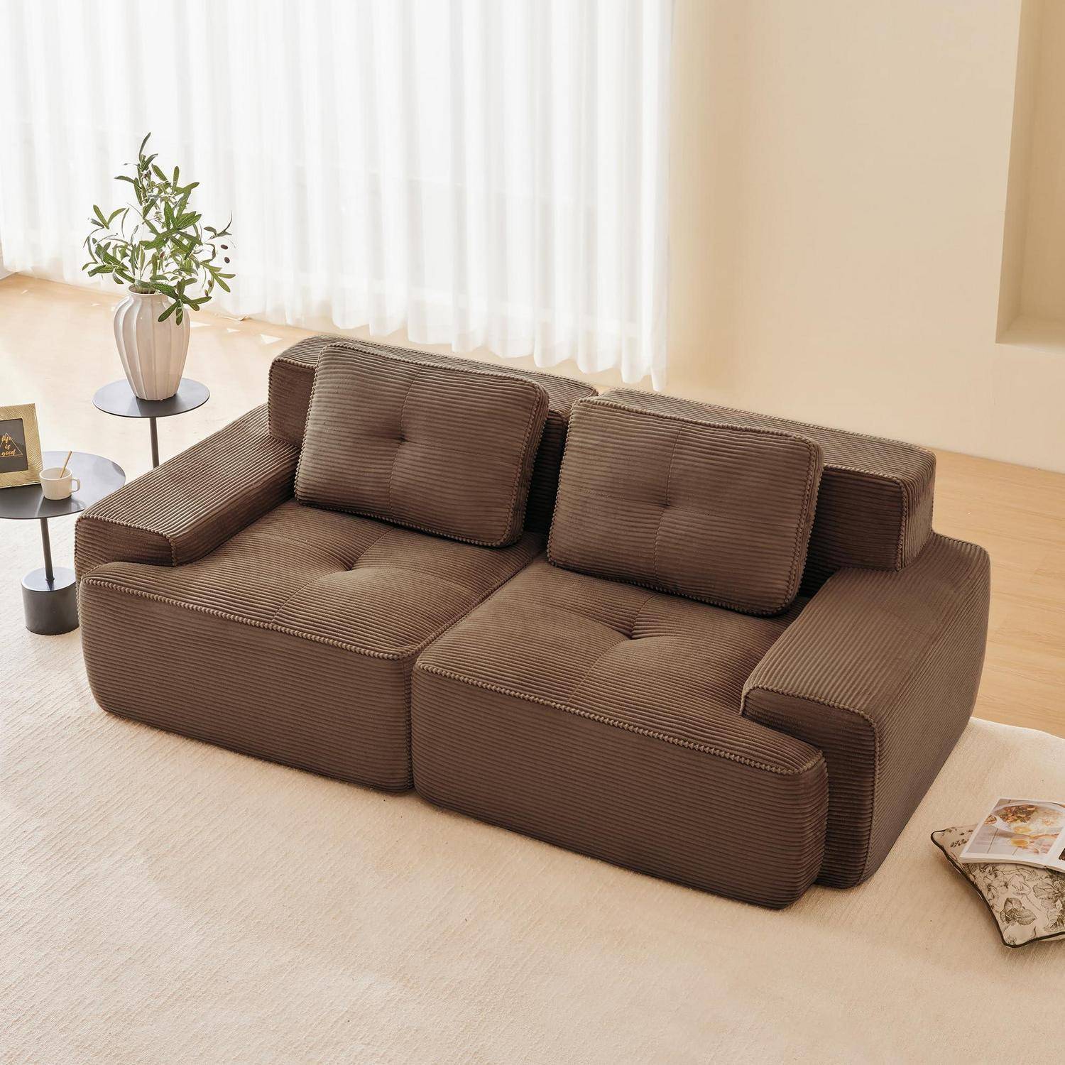 Braunes Cordsofá mit zwei Kissen in einem hellen, minimalistischen Wohnzimmer, neben einem Beistelltisch mit einer Pflanze und einem Buch auf dem Boden.