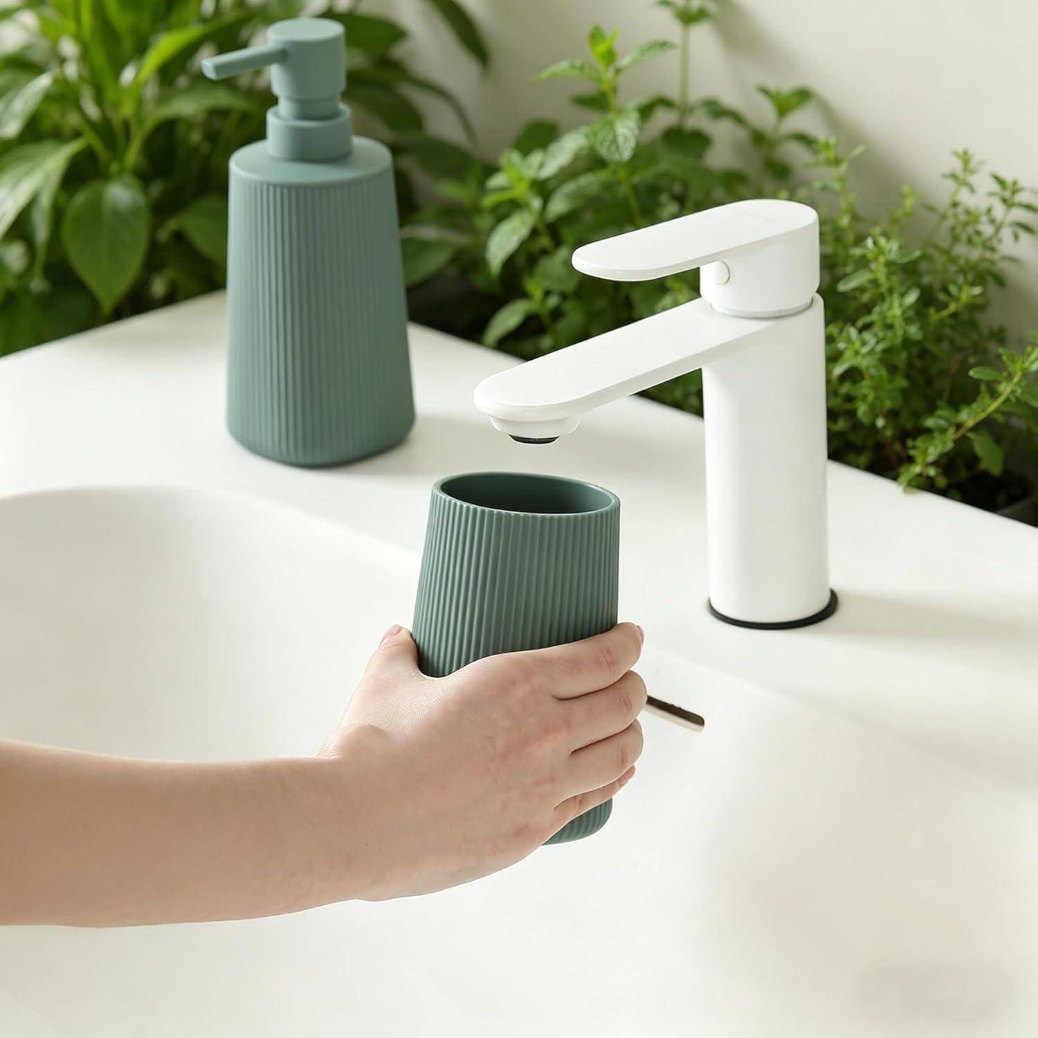 Eine Hand hält einen grünen gerippten Becher unter einen weißen Badezimmerhahn an einem Waschbecken, mit einem passenden Seifenspender und Grünpflanzen im Hintergrund.