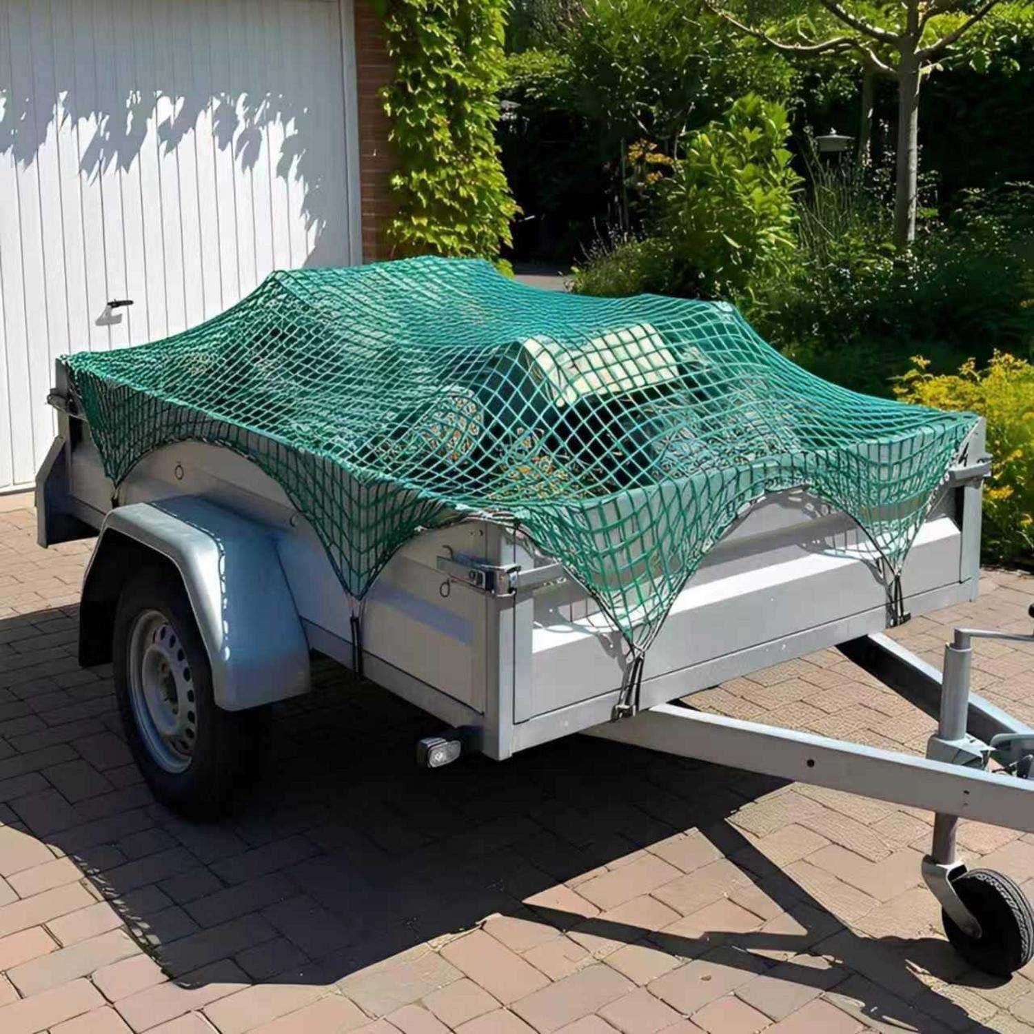 Kleiner Nutzanhänger mit grünem Ladungsnetz bedeckt, auf einer Ziegelpflaster-Einfahrt neben einem weißen Gebäude und Grünflächen geparkt.