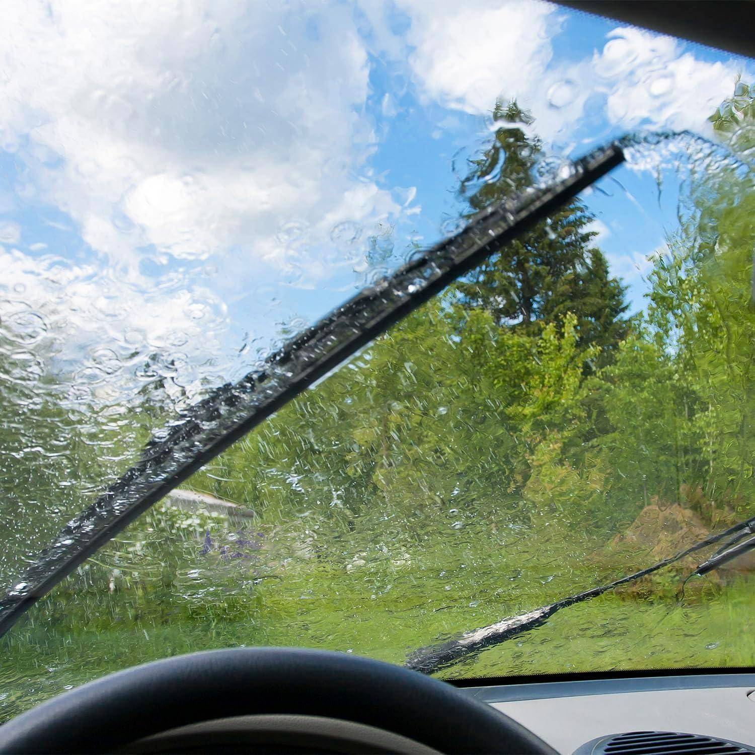 Aussicht von innen aus einem Auto durch eine regenverschmierte Windschutzscheibe, mit Scheibenwischern, die eine grüne bewaldete Landschaft und einen bewölkten Himmel freimachen.