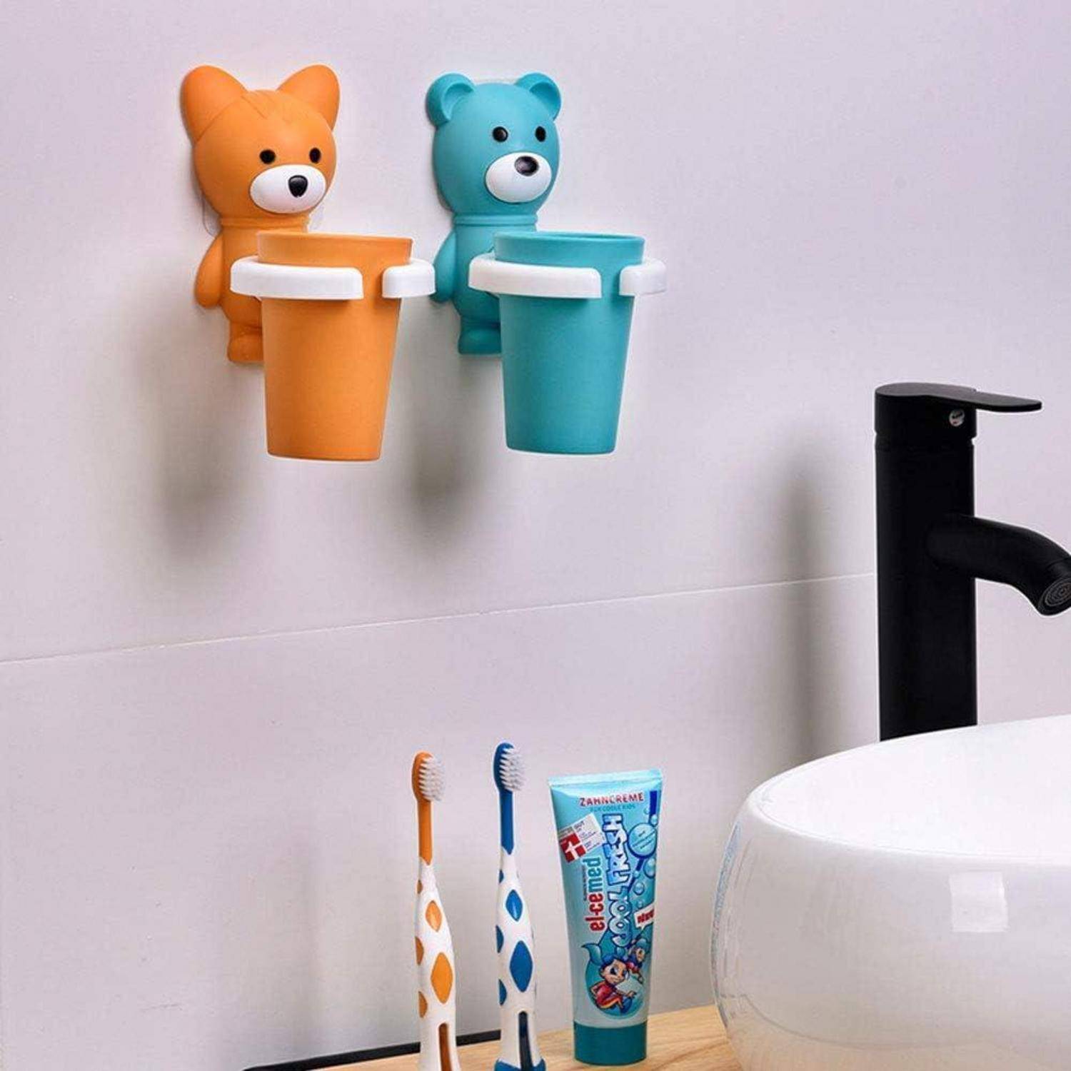 Zwei bärenförmige Zahnbürstenhalter an der Wand, einer orange und einer blau, mit einer Zahnpastatube und Zahnbürsten neben einem Badezimmerspülbecken und einem Wasserhahn.