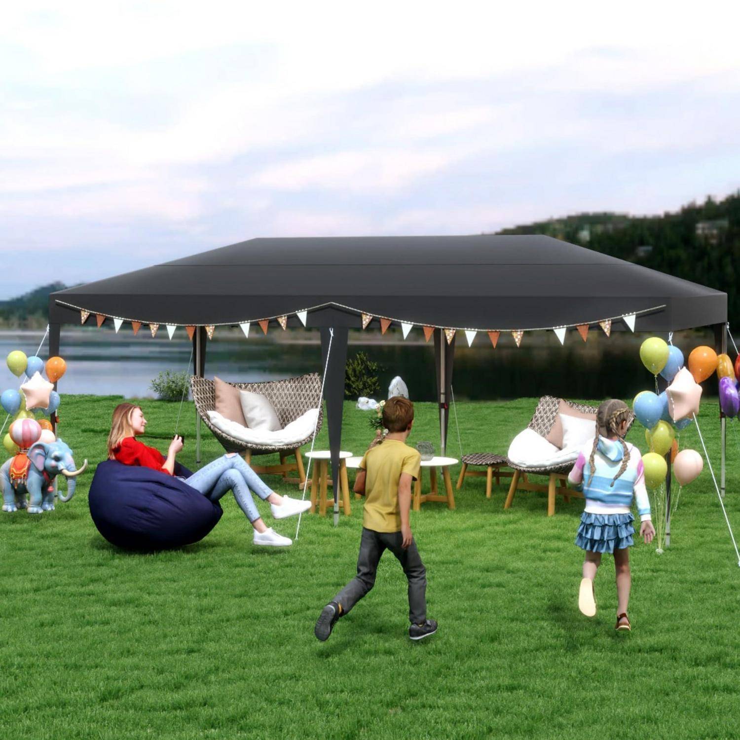 Kinder spielen in der Nähe eines schwarzen Pavillonzeltes auf einer Wiese am Wasser, mit Luftballons, Wimpelketten, Sitzsäcken und hängenden Stühlen bei einer festlichen Outdoor-Veranstaltung.