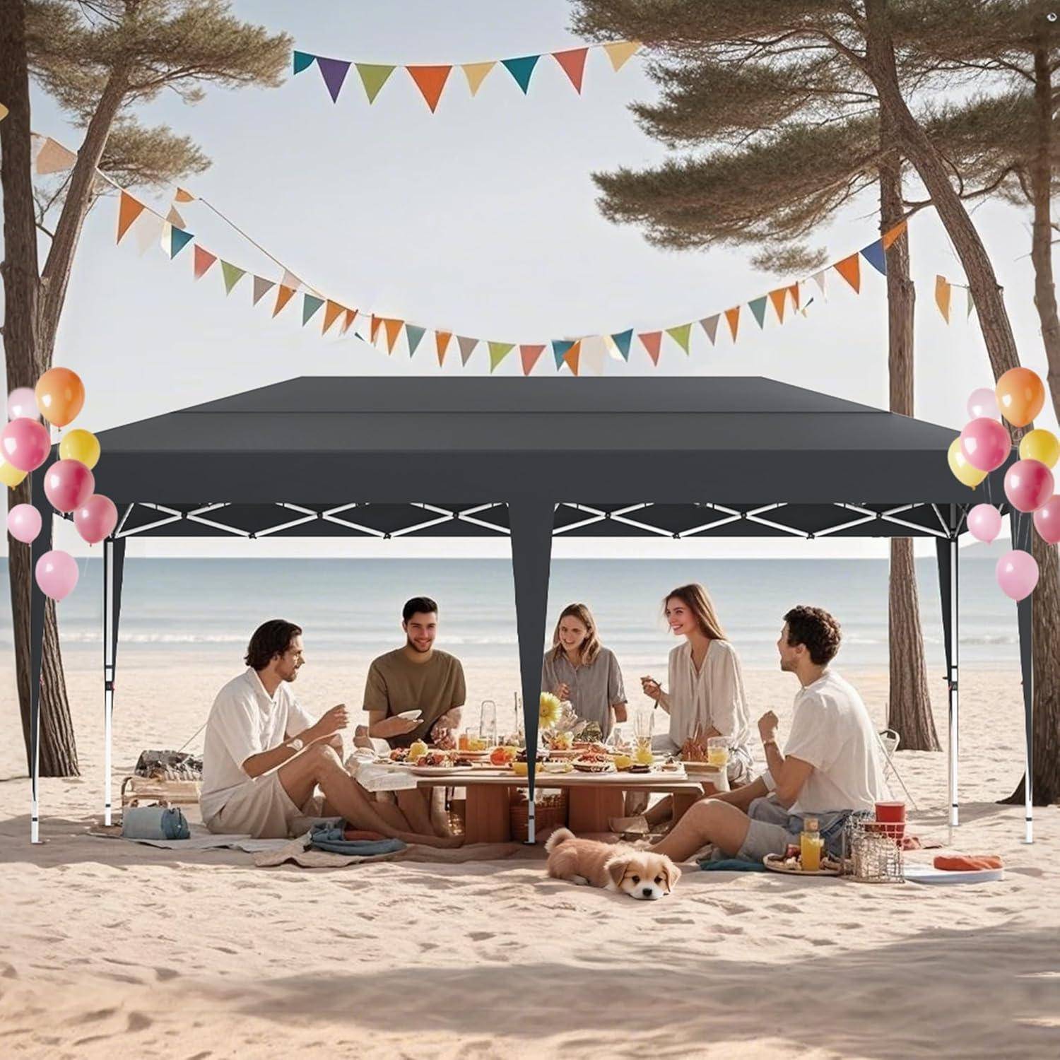 Freunde teilen ein Strandpicknick unter einem schwarzen Baldachin mit Wimpelketten und Luftballons, sitzend um einen niedrigen Tisch mit einem Hund im Sand am Meer.