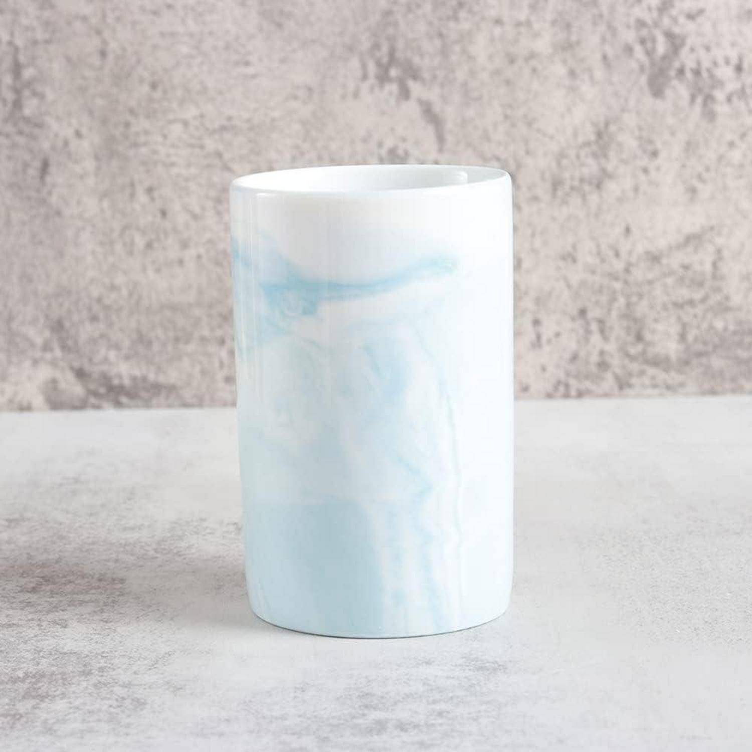Weiße zylindrische Tasse mit subtlem hellblauem Marmorierungsmuster auf einer grauen Oberfläche vor einem strukturierten grauen Hintergrund.