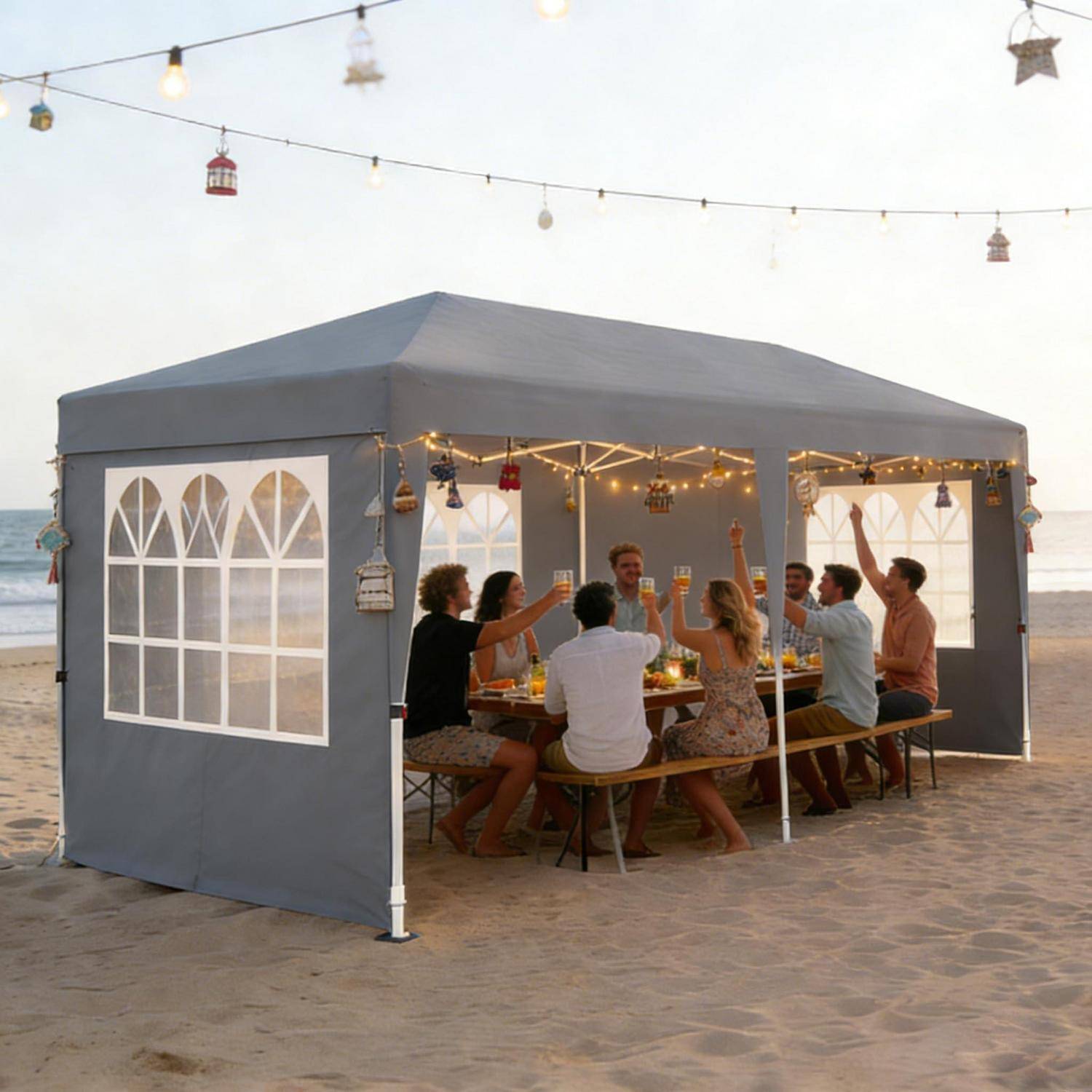 Menschen stoßen an einem Esstisch in einem grauen Strandzelt an, das bei Sonnenuntergang mit Lichtern und Laternen dekoriert ist.