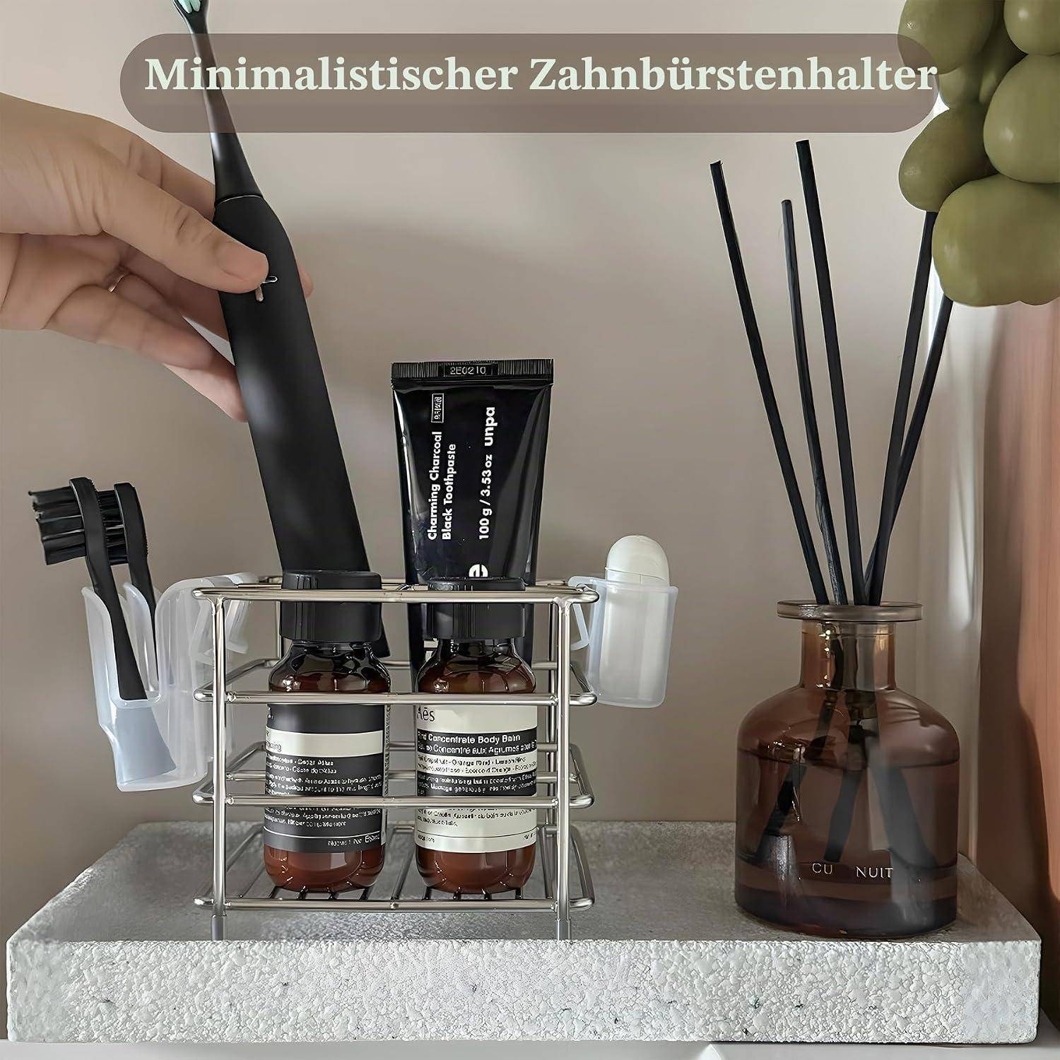 Eine Hand legt eine elektrische Zahnbürste in einen minimalistischen Badezimmer-Organizer, der Zahnpasta, Hautpflageflaschen und Bürstenhalter neben einem Raumduft-Diffuser mit Holzstäbchen enthält.