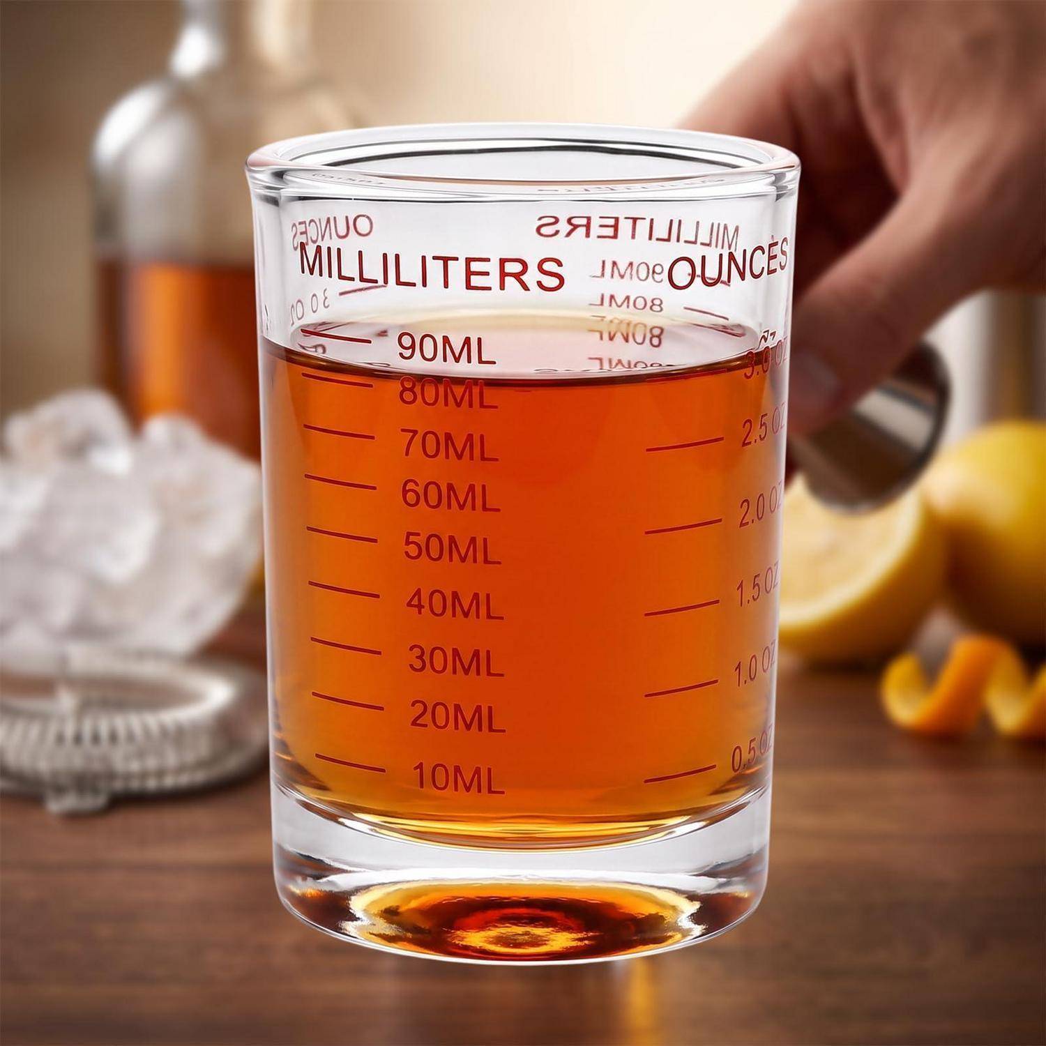 Glaskupfer mit bernsteinfarbener Flüssigkeit, gekennzeichnet in Millilitern und Unzen bis zu 90 ml/3 oz, gehalten von einer Hand auf einer Arbeitsplatte in der Küche.