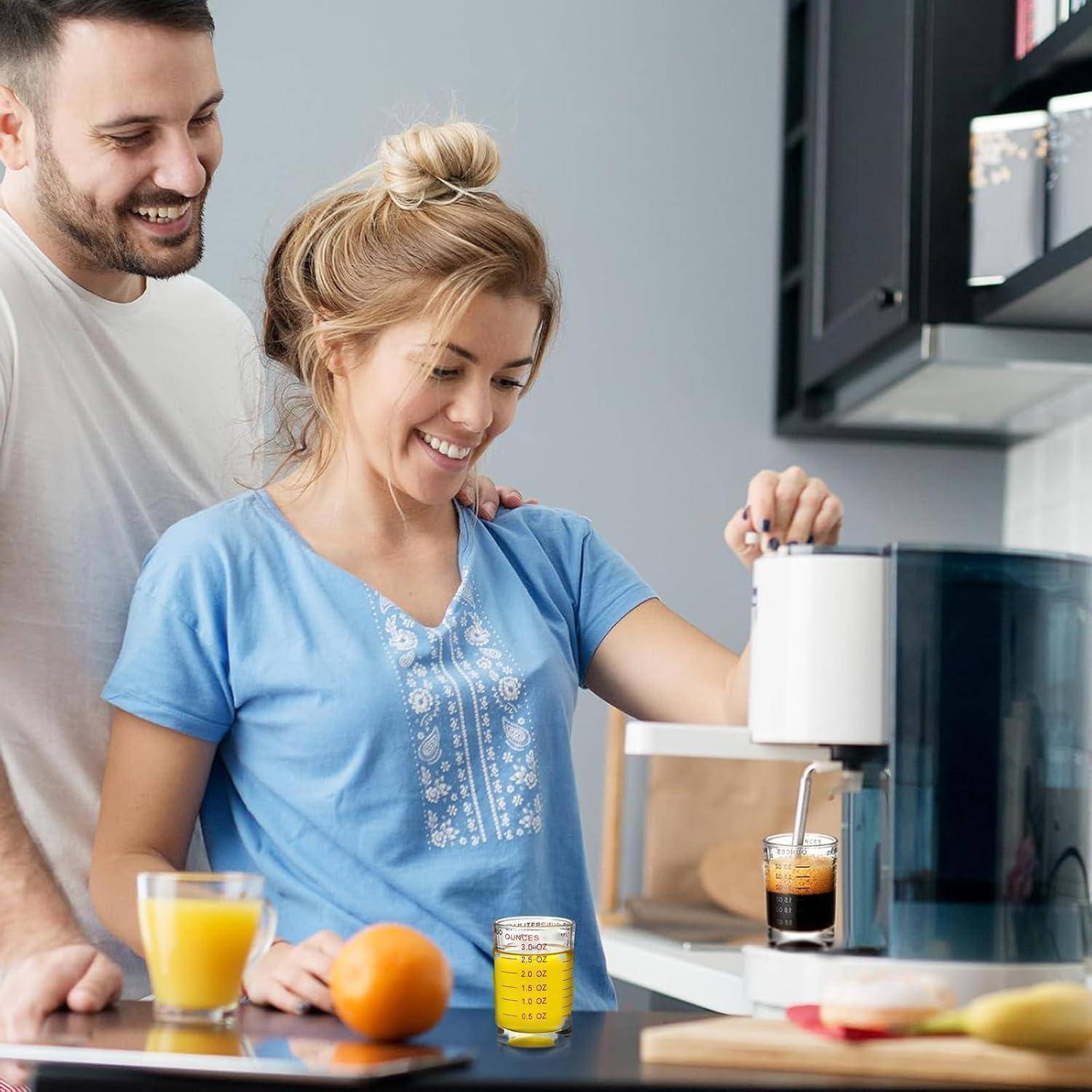 Lächelnde Frau, die eine Kaffeemaschine benutzt, während ein Mann in einer Küche zuschaut, mit Saft und Obst auf der Arbeitsplatte.