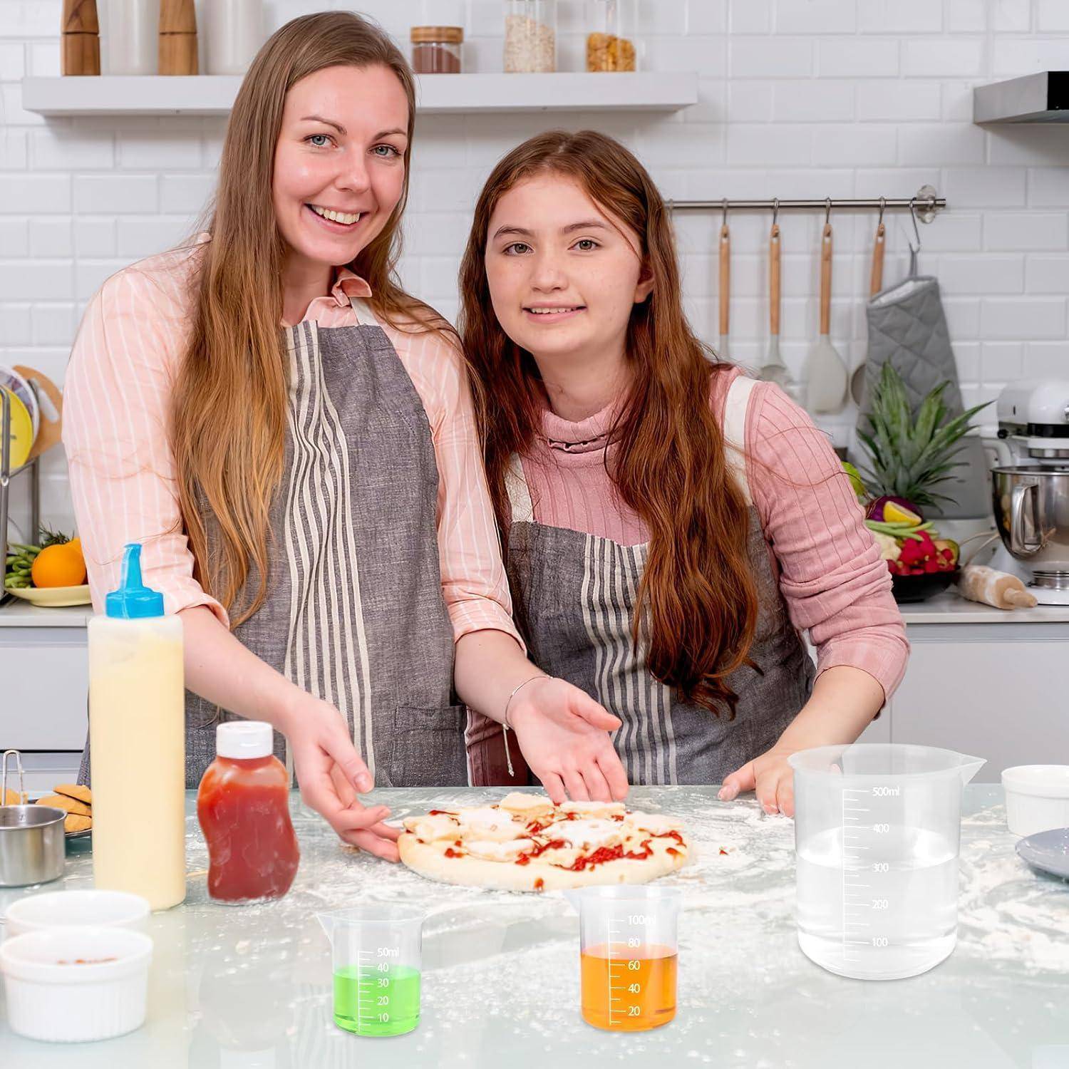 Zwei Frauen in Schürzen lächeln, während sie Pizzateig in einer Heimatküche zubereiten, mit Mehl, Messbechern und Würzmitteln auf der Arbeitsplatte.