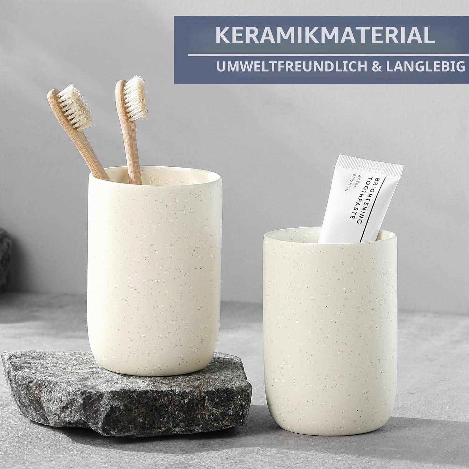 Zwei cremefarbene Keramik-Badezimmertassen auf einer Steinplatte, die eine Zahnbürsten hält und die andere eine Zahnpastatube, mit deutschem Text „KERAMIKMATERIAL