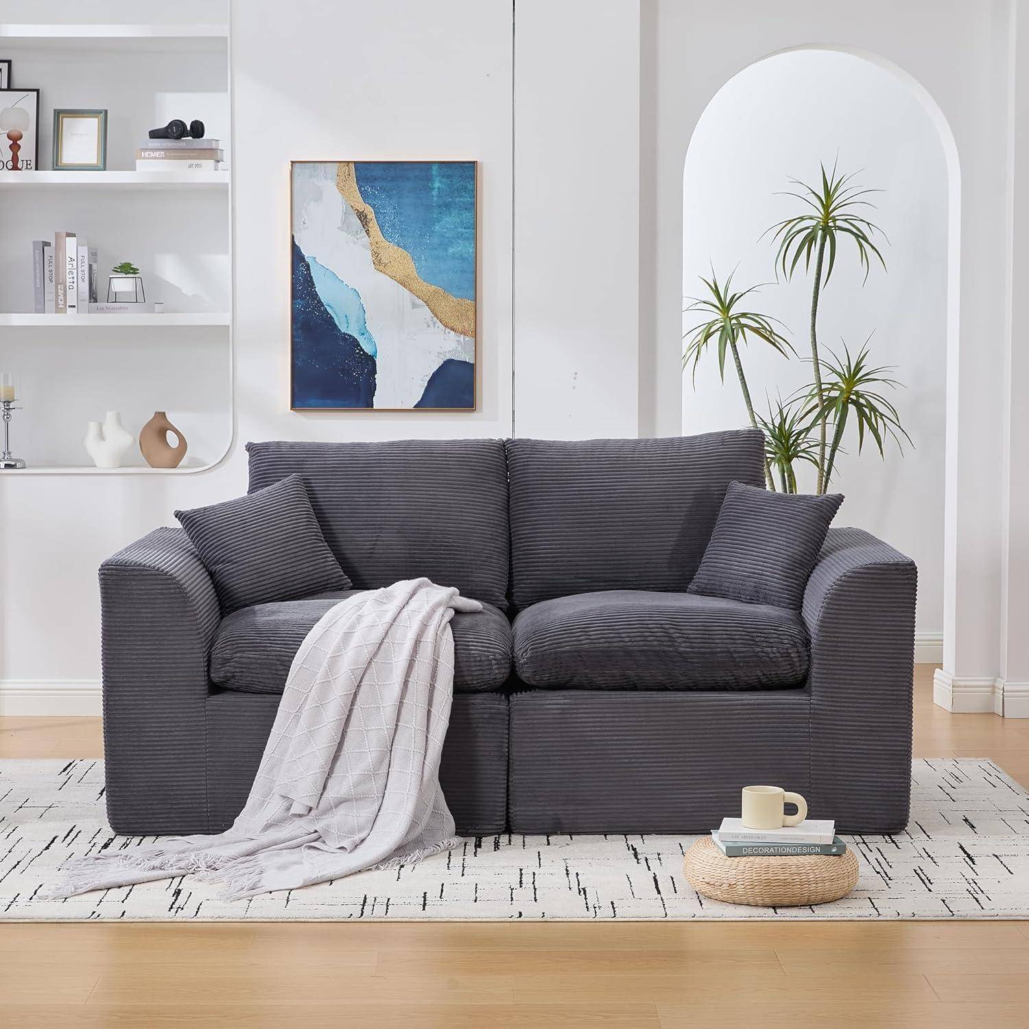Modernes Wohnzimmer mit dunkelgrauem Sofa, Wurfdecke, abstrakter Wandkunst, Regalen, Topfpflanze und kleinem Kaffee-Set auf hellem Teppich.