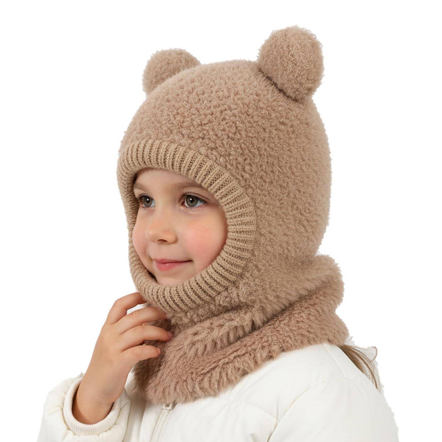 Kleines Kind in einer beigen gestrickten Balaklava mit Bärenohren und Schal, weiße Jacke tragend, in Profilansicht vor weißem Hintergrund posiert.