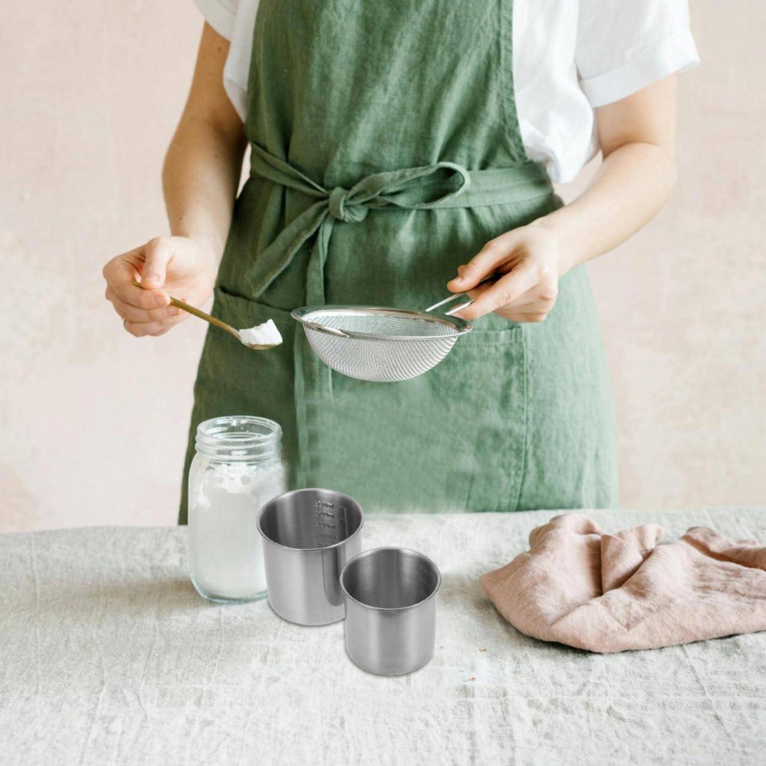 Person in grüner Schürze, die ein weißes Pulver in eine Schüssel neben Gläsern und Messbechern auf einem Küchentisch siebt.