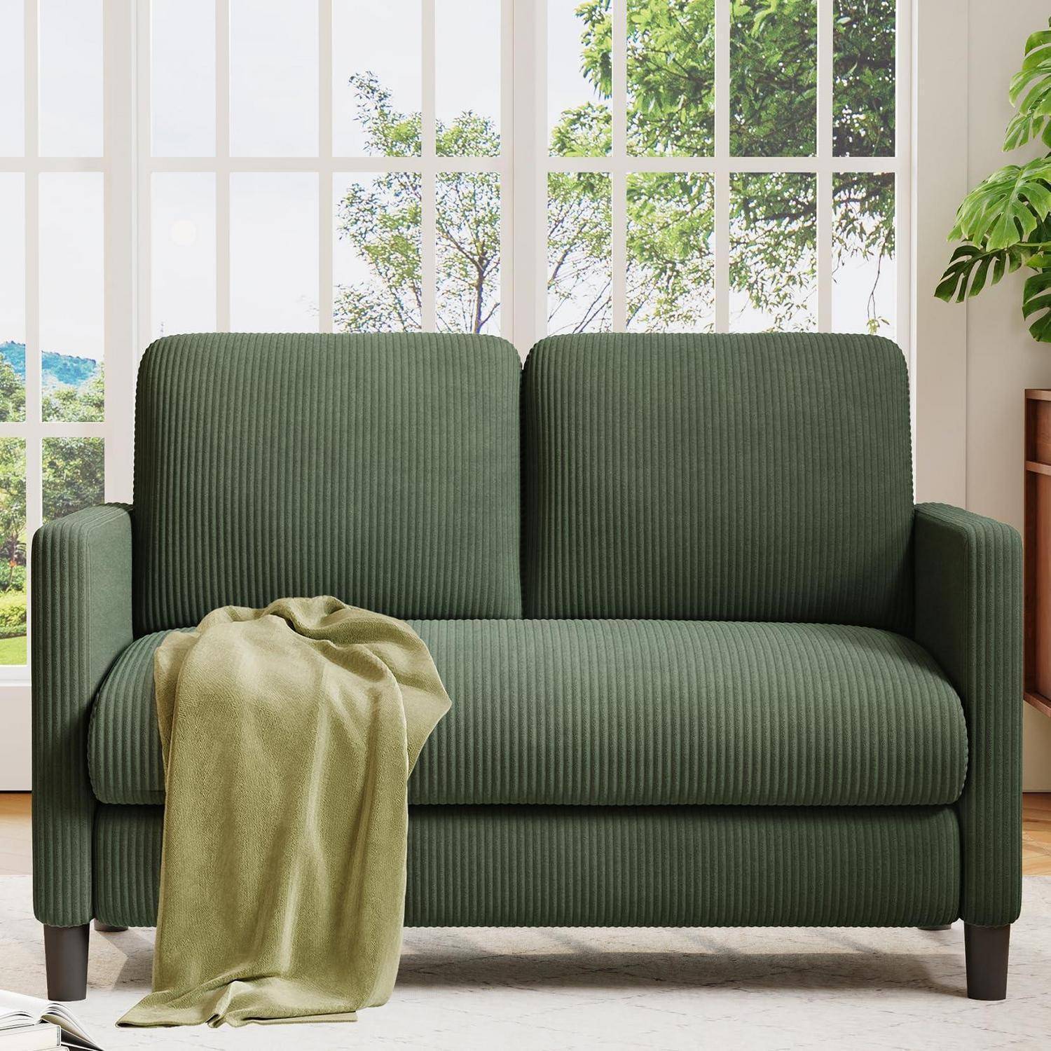 Grünes geripptes Zweisitzer-Sofa mit einer drapiertem helloliven Plaid in einem hellen Wohnzimmer neben hohen Fenstern und Grünflächen im Freien.