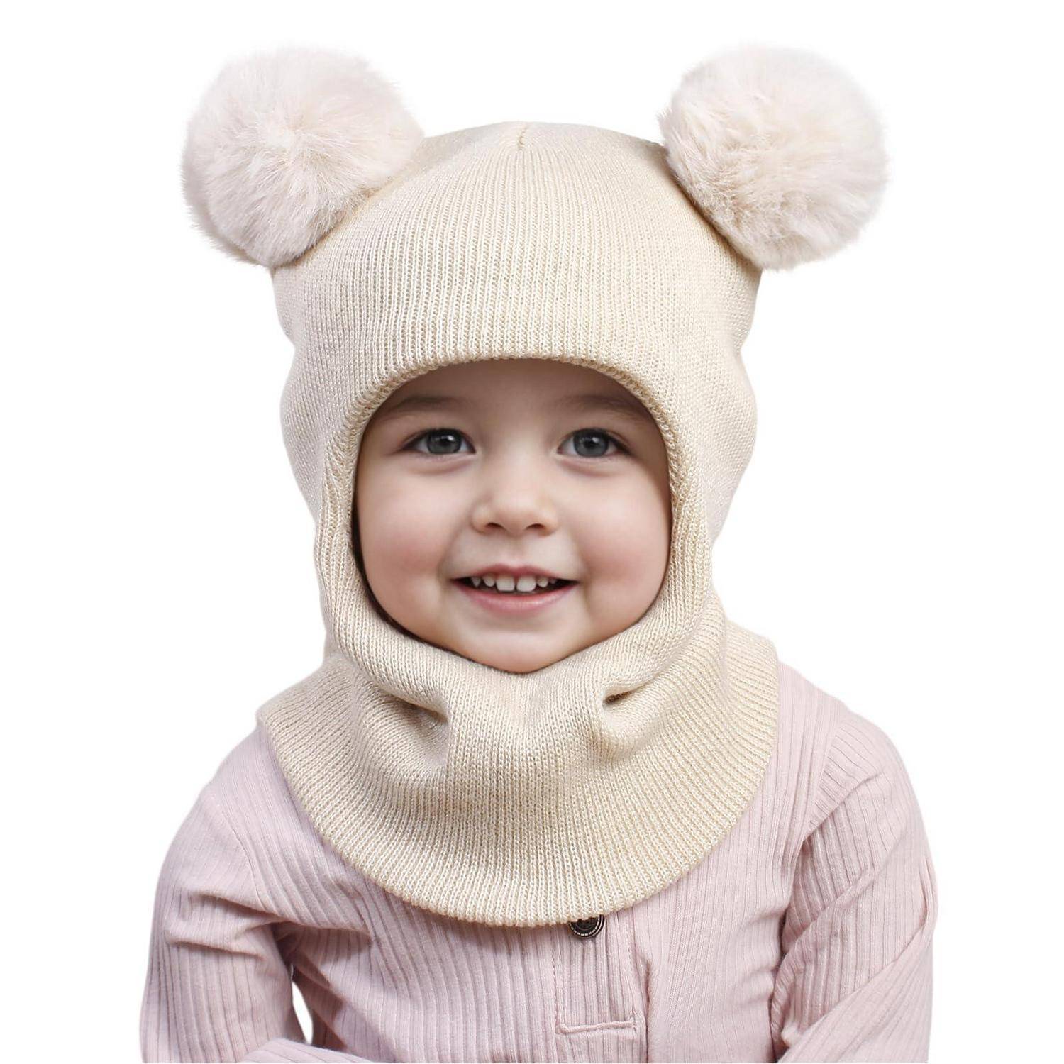 Lachendes Baby mit einer cremefarbenen Strickmütze mit Bärenohren und passendem Halstuch, in einem hellen rosa Outfit auf weißem Hintergrund.