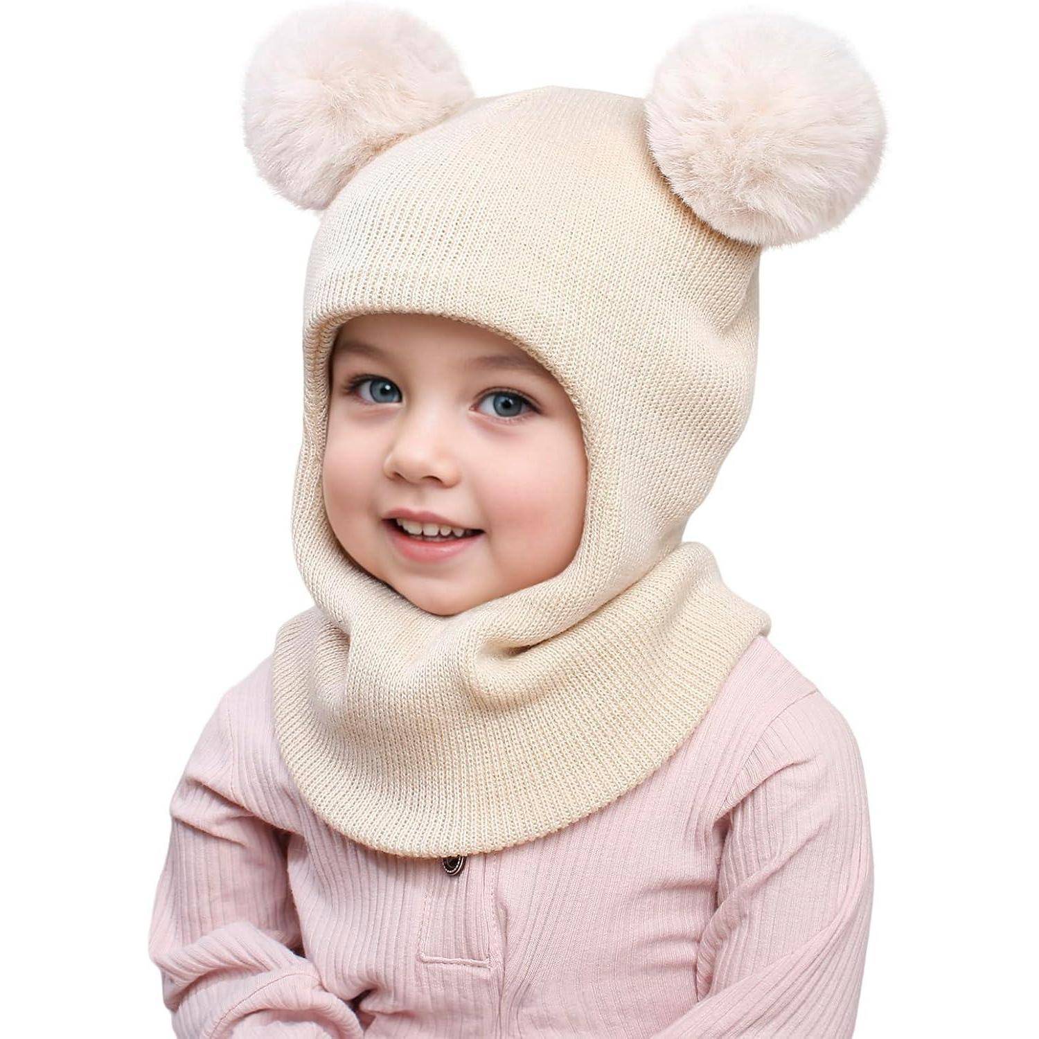 Lachendes Kleinkind, das eine cremefarbene Strickbalaclavas mit Pompon-Ohren und ein blasses rosafarbenes Hemd vor weißem Hintergrund trägt.