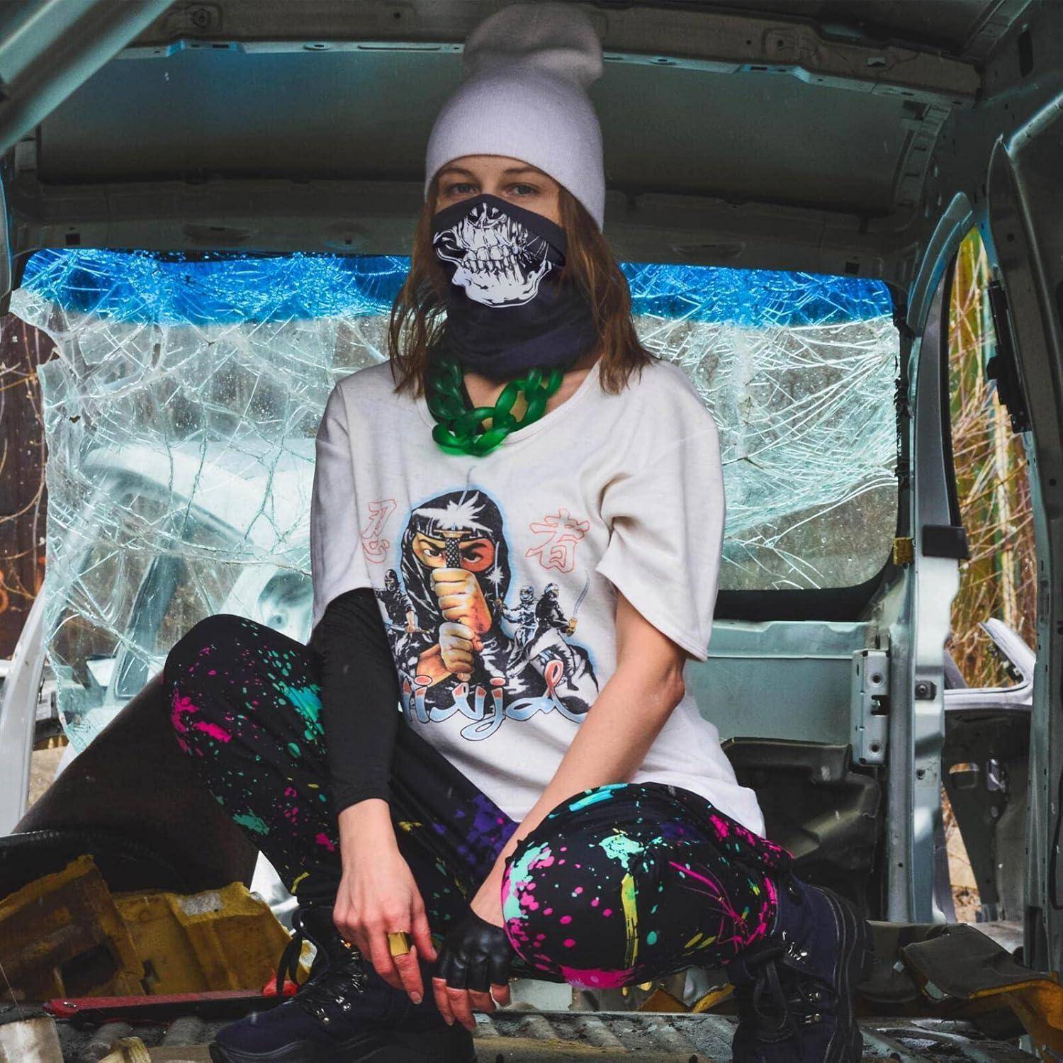 Eine maskierte Person in einer Wollmütze sitzt in einem beschädigten Lieferwagen mit zersplitterten Fenstern, trägt ein bedrucktes T-Shirt und mit Farbe bespritzte Hosen.