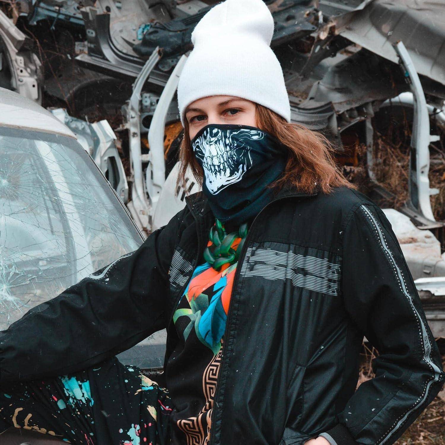 Eine Person in einer weißen Beanie und Gesichtsbedeckung steht neben einem beschädigten Auto in einer Szene industrieller Trümmer.
