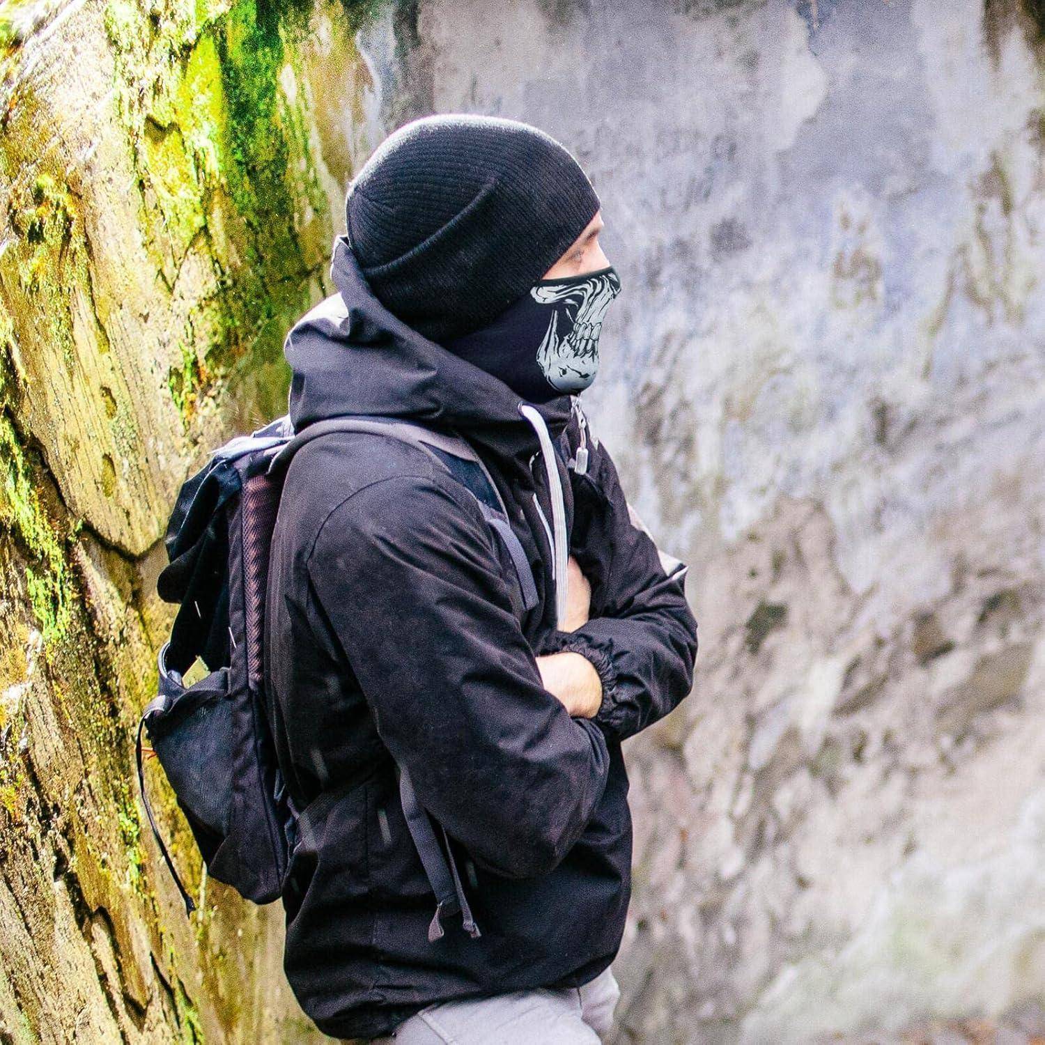 Maskierter Wanderer in schwarzer Jacke und Mütze mit Rucksack, neben einer moosbedeckten Steinmauer im Freien stehend.