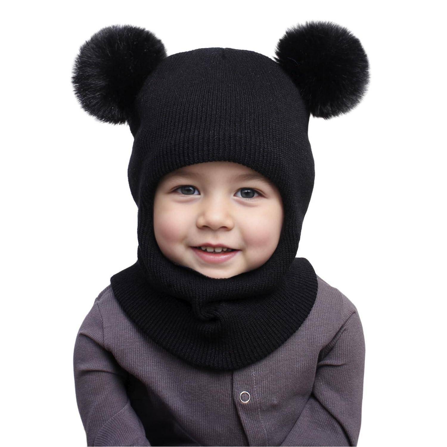 Lachendes Baby, das eine schwarze gestrickte Mütze mit zwei Bommel-Ohren und einer passenden Gesichtsmaske trägt, vor weißem Hintergrund.