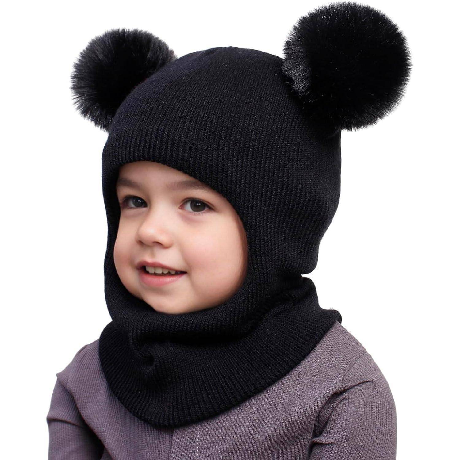 Junges Kind, das eine schwarze Strickmütze mit zwei flauschigen Bommel und Gesichtsöffnung trägt, vor weißem Hintergrund gezeigt.