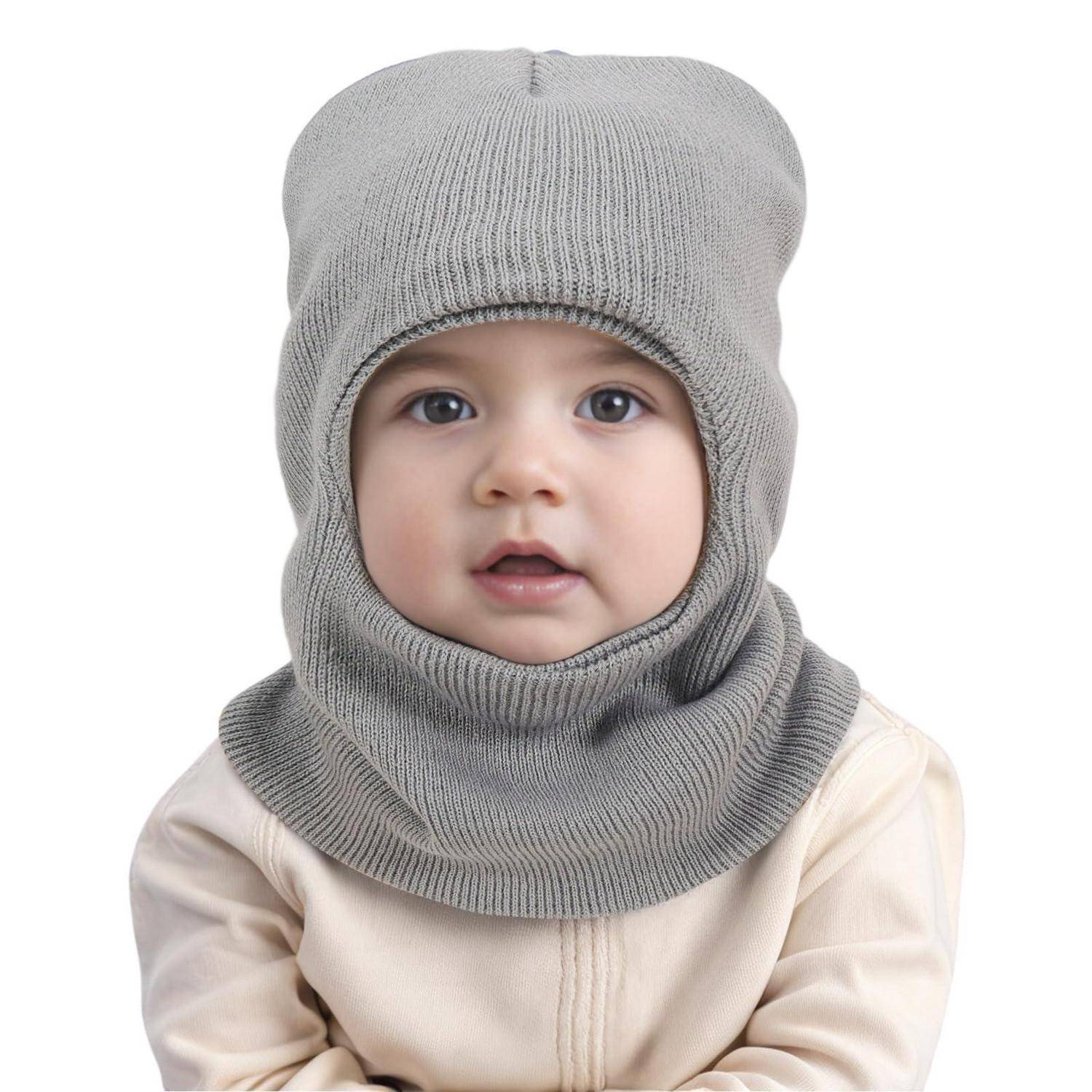 Ein Baby mit einer grauen Strickbalaclava und beigefarbenem Outfit, frontal vor weißem Hintergrund.