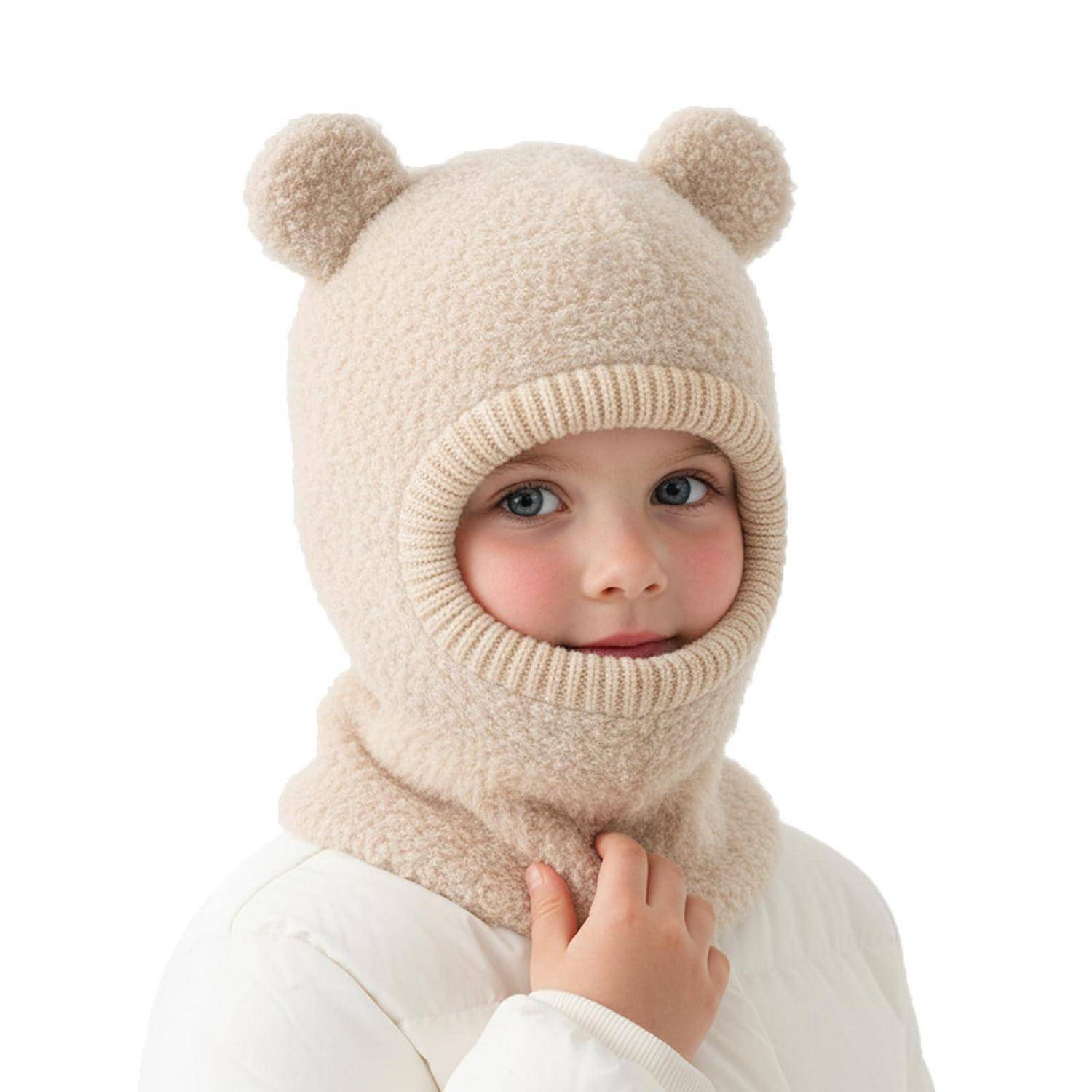 Kleines Kind, das eine cremefarbene Teddybär-Winterbalaklava mit Ohrpuscheln trägt, blickt gegen einen weißen Hintergrund in die Kamera.
