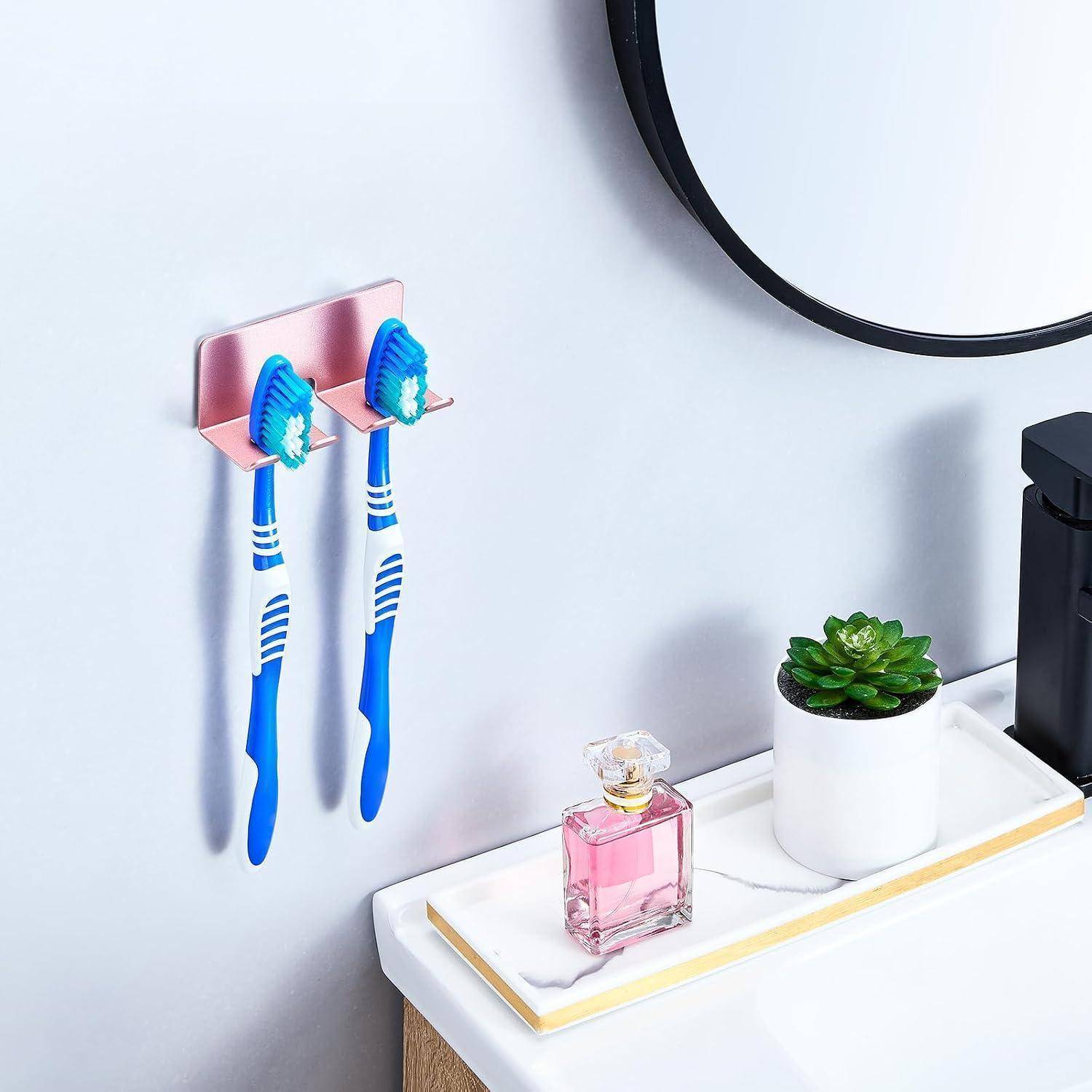 Zwei blaue Zahnbürsten hängen an einem rosa Wandhalter über einer Badezimmerablage mit einer Parfümflasche, einer kleinen Pflanze und einem Spiegel.
