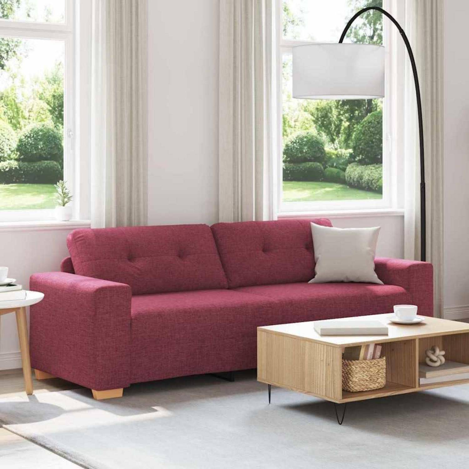 Ein magentafarbenes Sofa in einem hellen Wohnzimmer mit großen Fenstern, einer Stehlampe und einem hellem Holz-Couchtisch mit einem Tablett und einer Tasse.